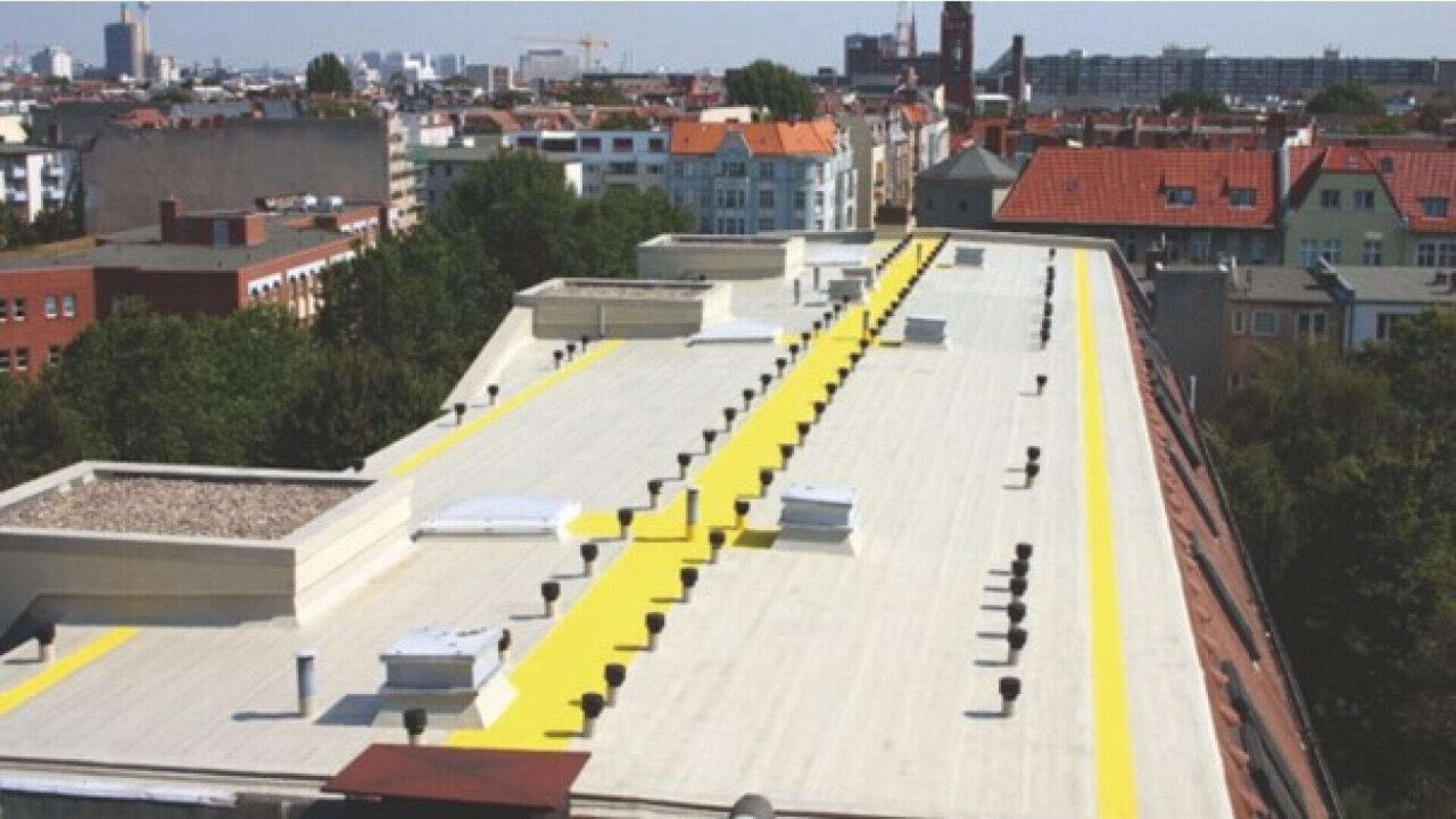 A flat rooftop on a city building features wide yellow safety paths running along its length, surrounded by various vents, skylights, and urban buildings in the background.