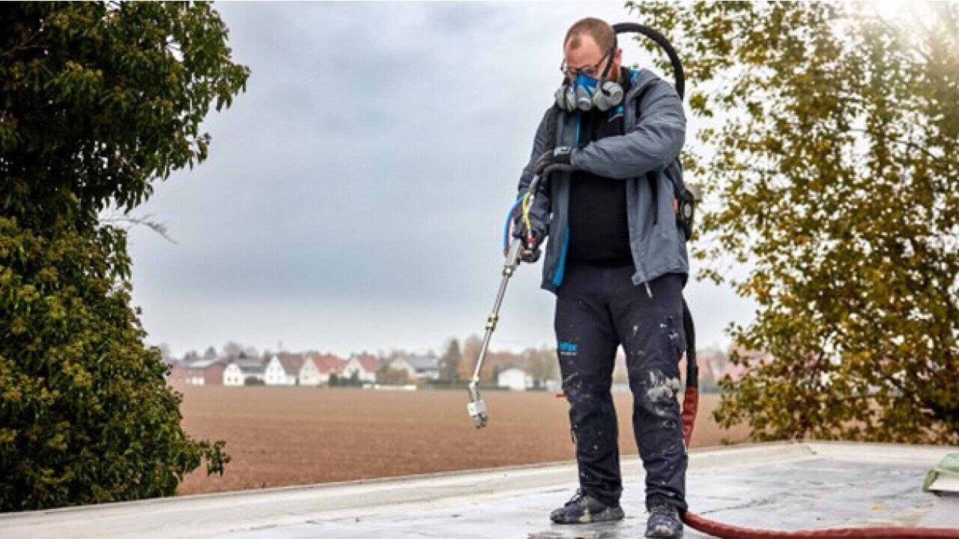 A person wearing protective gear and a respirator mask stands on a rooftop, operating spraying equipment, with trees and houses visible in the background under a cloudy sky.