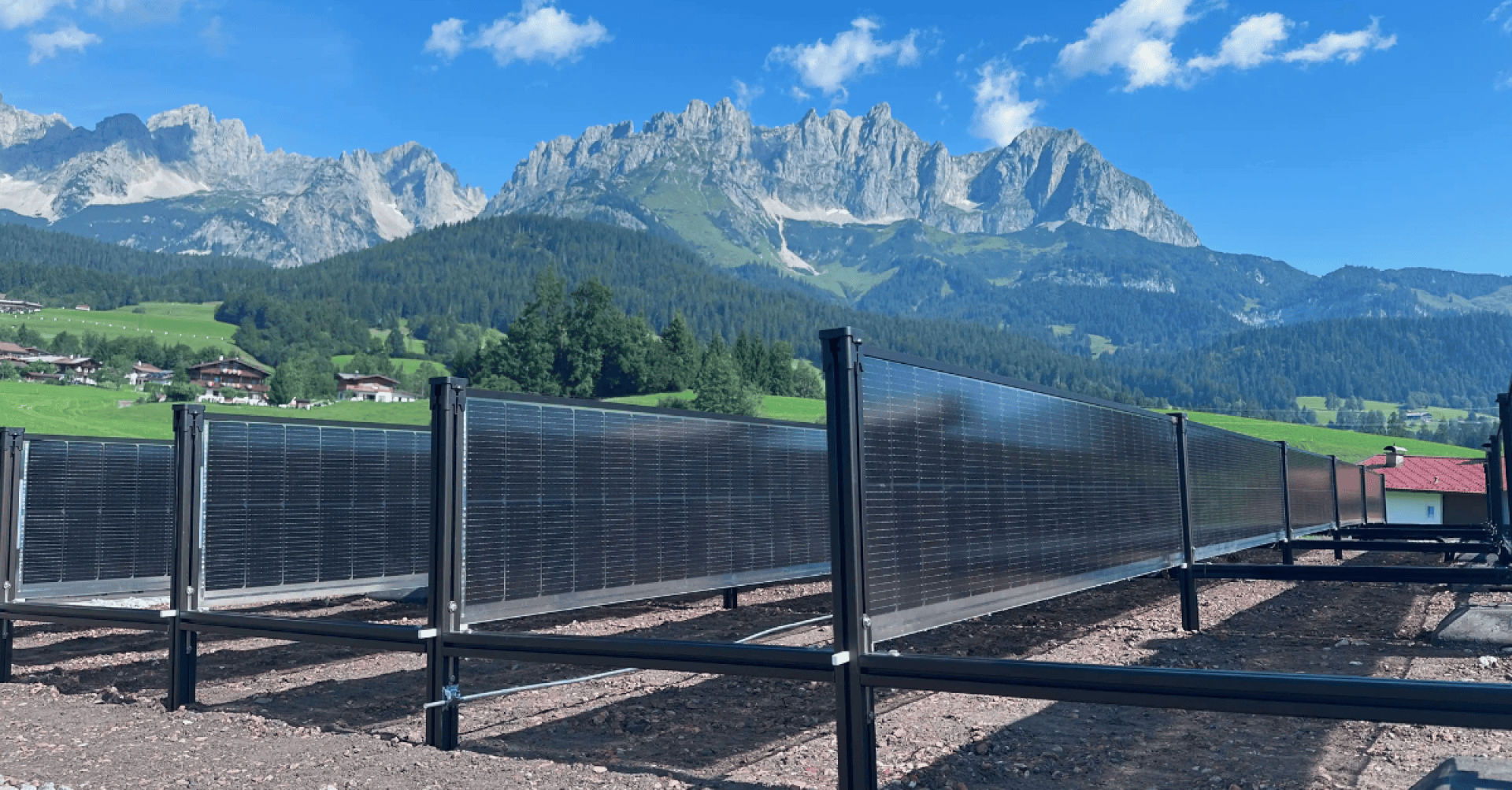Reihen von vertikalen Solarmodulen stehen auf einer Kiesfläche vor einer üppig grünen Landschaft mit Häusern, Bäumen und hohen, zerklüfteten Bergen unter einem strahlend blauen Himmel mit vereinzelten Wolken.