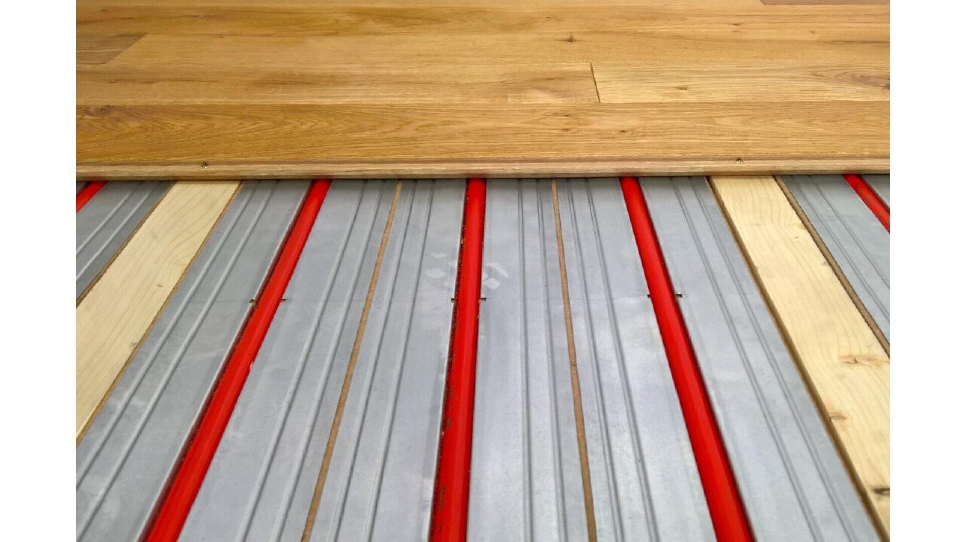 Close-up of an underfloor heating system with red pipes set in metal plates, partially covered by wooden floorboards.