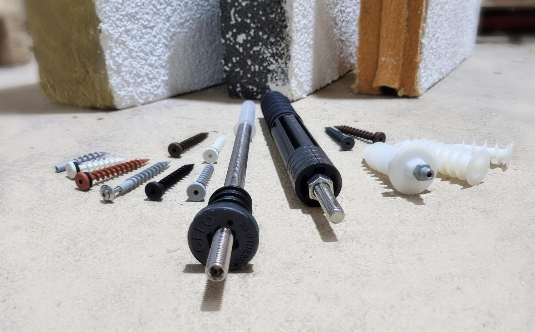 Various dowels, screws and anchors are displayed on a surface with three building material samples in the background, including concrete, brick and insulation boards.