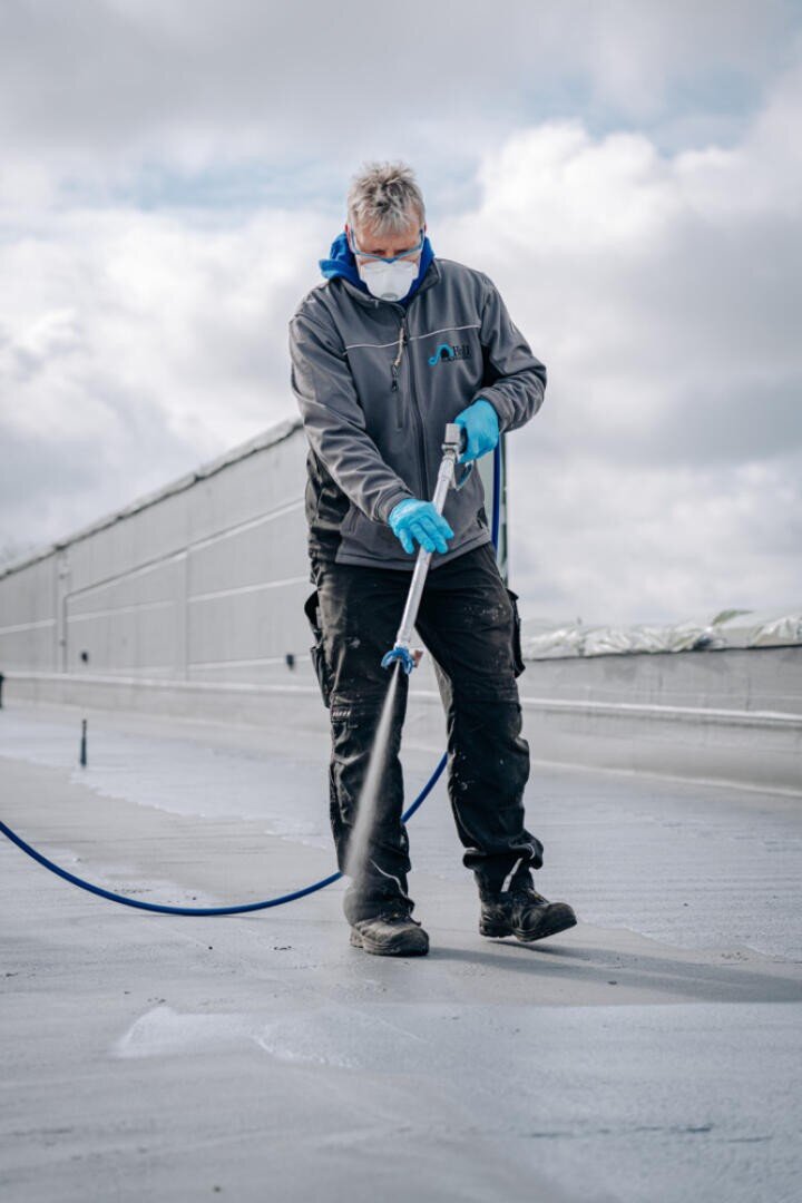 A person wearing a face mask, gloves and a gray jacket sprays a liquid onto a flat roof surface with a hose, while the sky is cloudy in the background.