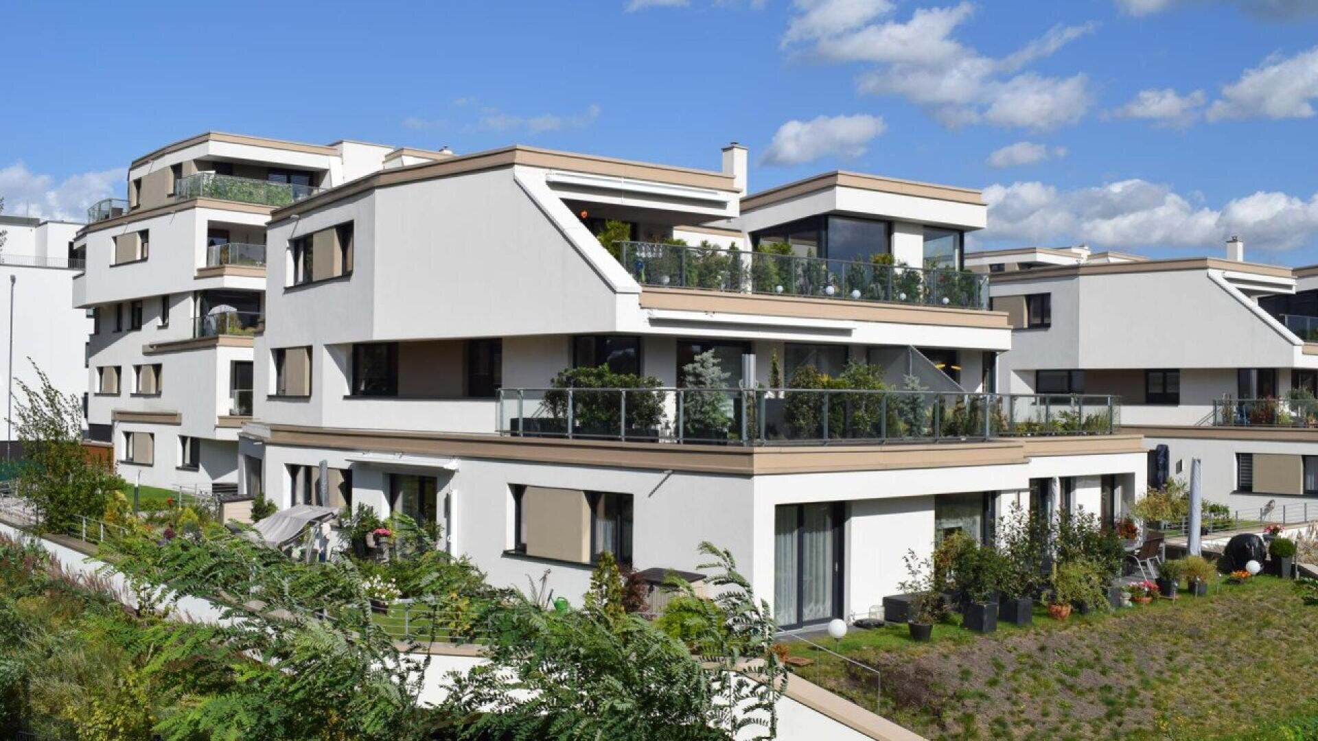 Modern white apartment buildings with large balconies and glass railings, surrounded by greenery and plants, under a partly cloudy blue sky.