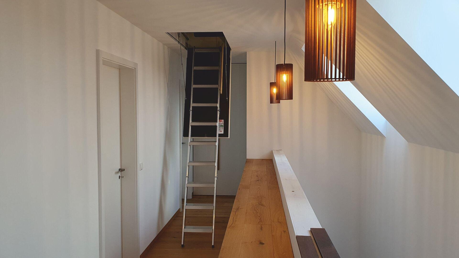 Ein moderner Flur mit weißen Wänden, Holzboden und drei Pendelleuchten. Eine ausziehbare Dachbodenleiter ragt von der Decke in der Nähe einer geschlossenen weißen Tür. Licht fällt durch schräge Fenster auf der rechten Seite ein.