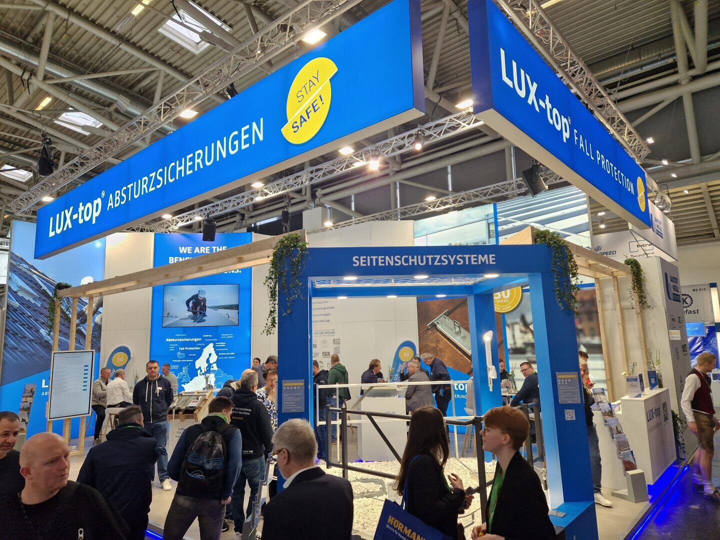 A busy exhibition stand for LUX-top, which offers fall protection systems, with people interacting with each other, banners with the company's branding and information displays under a large blue overhead sign.