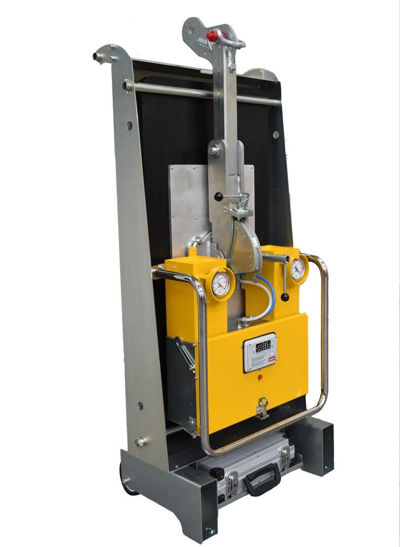 A vertical industrial lifting device with a yellow control box, pressure gauge, digital display, and metal frame, designed for handling heavy objects. The device stands on a sturdy base with wheels.