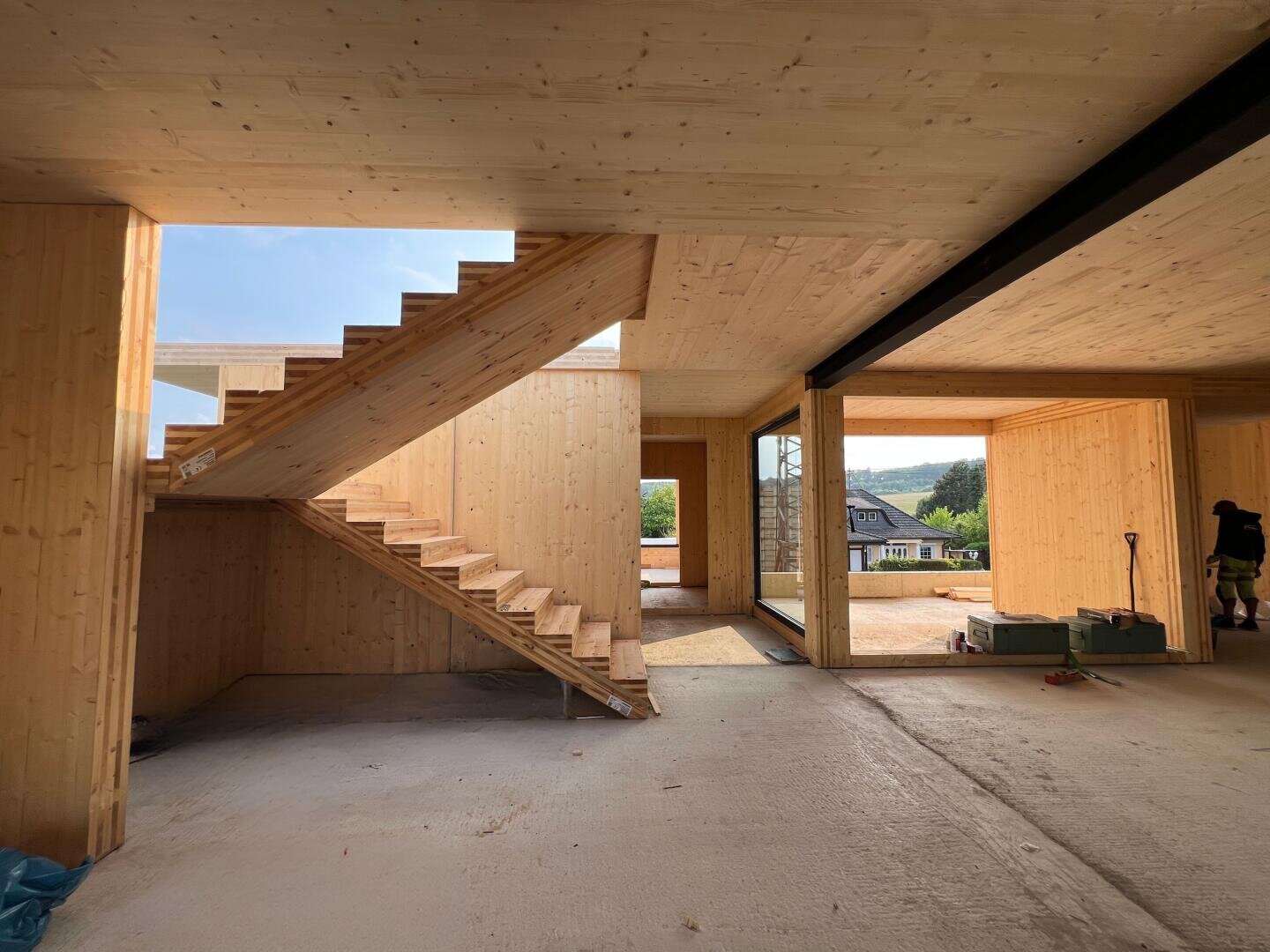 Innenansicht eines modernen, im Bau befindlichen Holzhauses mit unvollendetem Treppenhaus, freiliegenden Holzwänden, offenen Räumen und großen Fenstern, die natürliches Licht und einen Blick auf die Häuser und das Grün draußen bieten.
