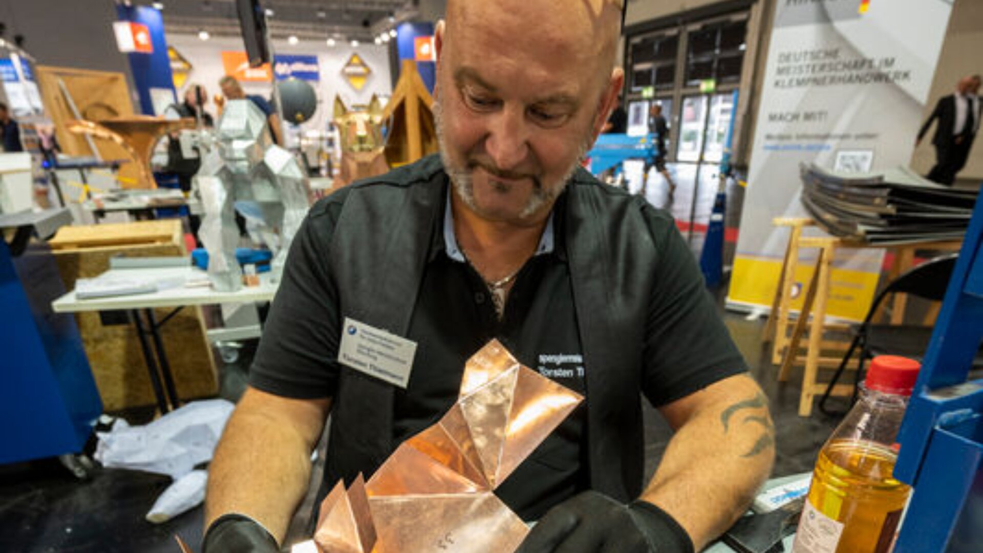 A man wearing black gloves works with a shiny, angular copper sculpture at a craft or trade show booth, surrounded by tools and informational displays.
