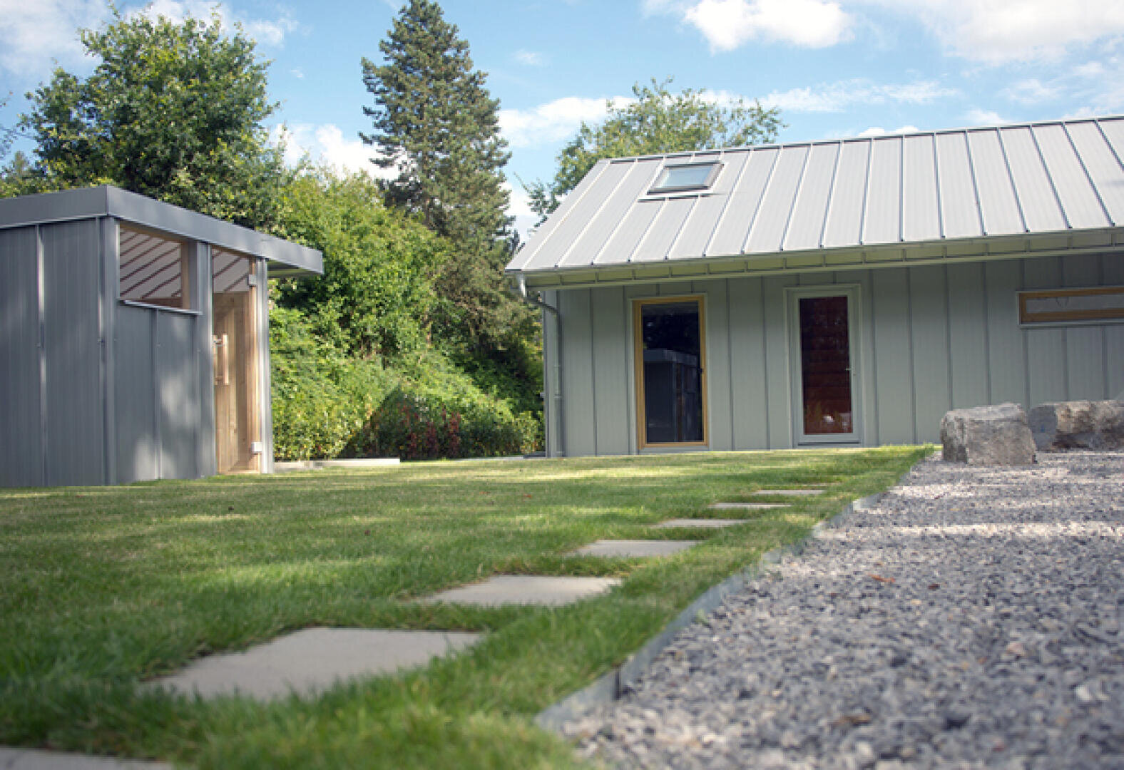 Modern backyard with green lawn, stone paved path, gravel area and two gray metal buildings with large windows, surrounded by trees under a blue sky.