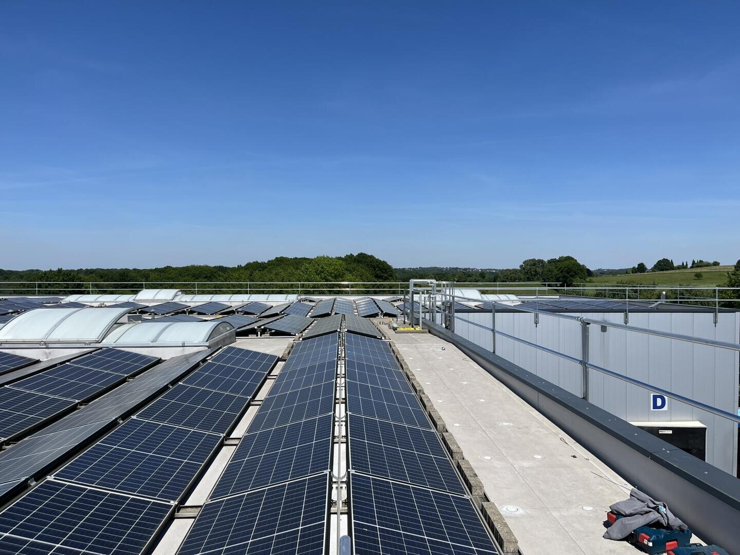 Reihen von Solarmodulen, die auf einem flachen Dach unter einem klaren blauen Himmel installiert sind, wobei im Hintergrund grüne Bäume und Felder zu sehen sind. Ein paar Kleidungsstücke oder Ausrüstungsgegenstände liegen auf dem Dach in der Nähe eines Geländers.
