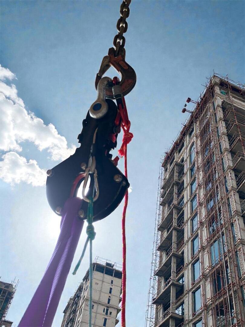 Eine Nahaufnahme eines schweren, an einer Kette aufgehängten Hakensystems mit bunten Seilen vor einem blauen Himmel und hohen, im Bau befindlichen Gebäuden mit Gerüsten.