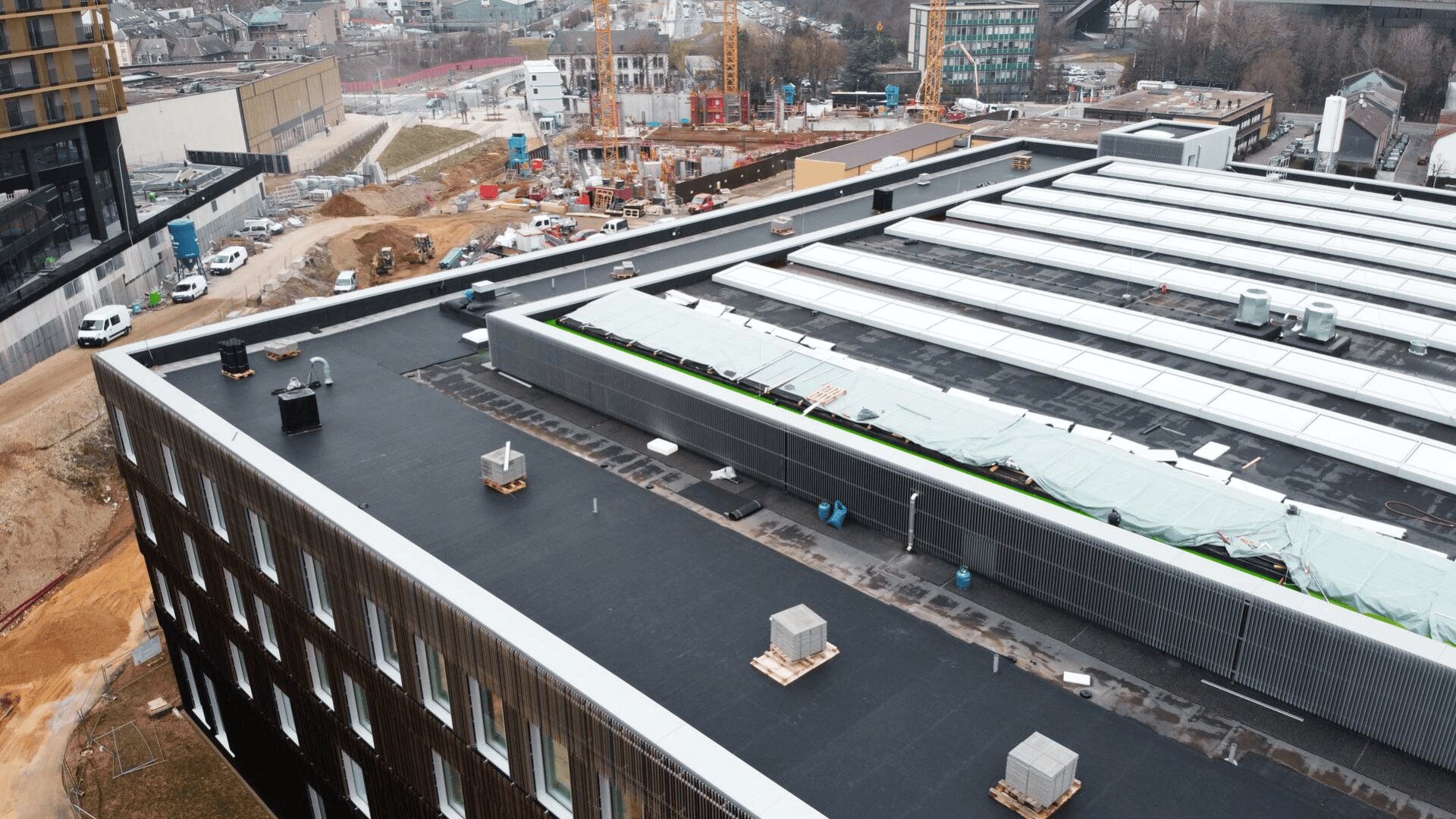 Luftaufnahme einer Baustelle mit Kränen, Geräten und teilweise errichteten Gebäuden. Im Vordergrund befindet sich ein großes Gebäude mit Flachdach, Lüftungsöffnungen und überdachten Bereichen. Im Hintergrund sind Wege und nahe gelegene Gebäude zu sehen.