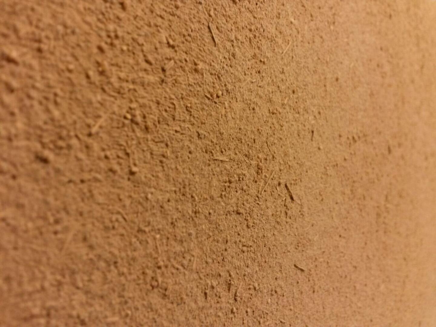 Nahaufnahme einer strukturierten braunen Oberfläche, möglicherweise einer Wand oder eines Stücks Pappe, mit rauen, ungleichmäßigen Fasern und kleinen Partikeln. Die Beleuchtung hebt die Textur und Tiefe des Materials hervor.