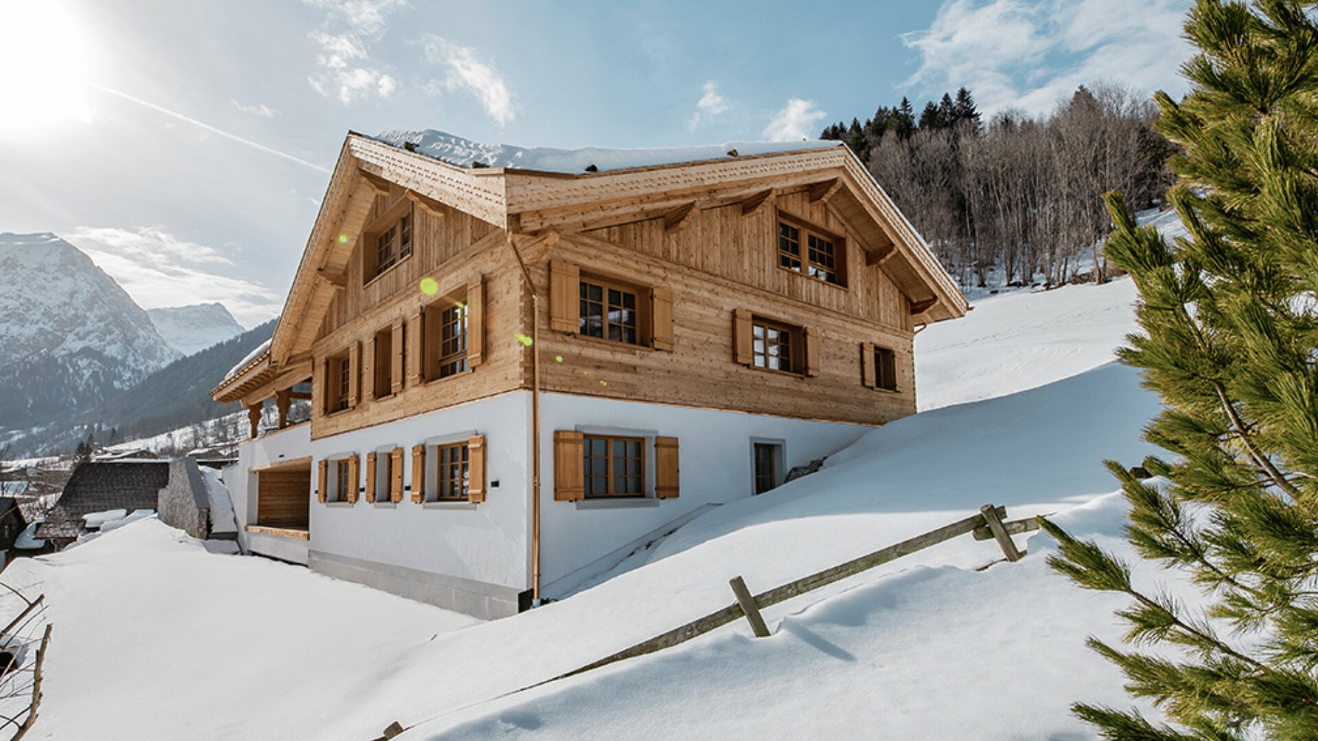 Ein Holzchalet mit weißen unteren Wänden und Fensterläden liegt auf einem schneebedeckten Hügel mit Kiefern und Bergen im Hintergrund unter einem blauen Himmel.