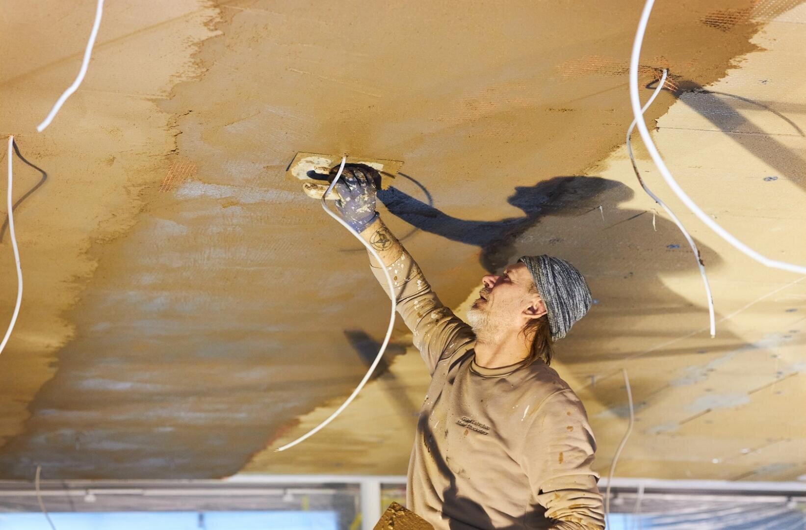 Ein Bauarbeiter mit grauer Mütze und braunem Hemd glättet mit einer Kelle den Putz an einer Decke, umgeben von herabhängenden Stromkabeln.