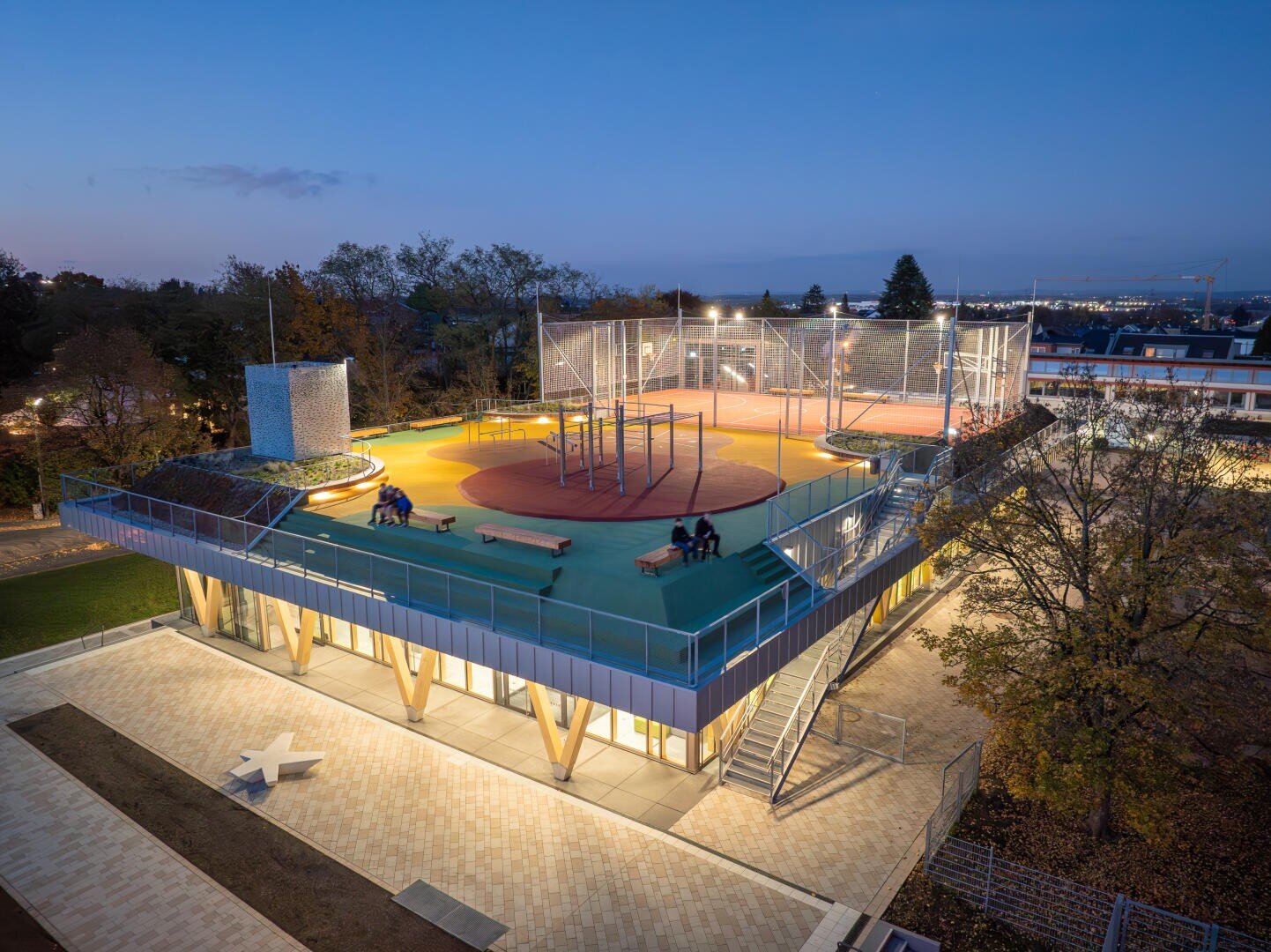 Ein beleuchtetes modernes Gebäude in der Abenddämmerung mit einem Sportplatz auf dem Dach mit buntem Bodenbelag, Zäunen, Bänken und Menschen, die dort sitzen. Die untere Ebene ist mit Glaswänden versehen, und Bäume umgeben das Gebäude.