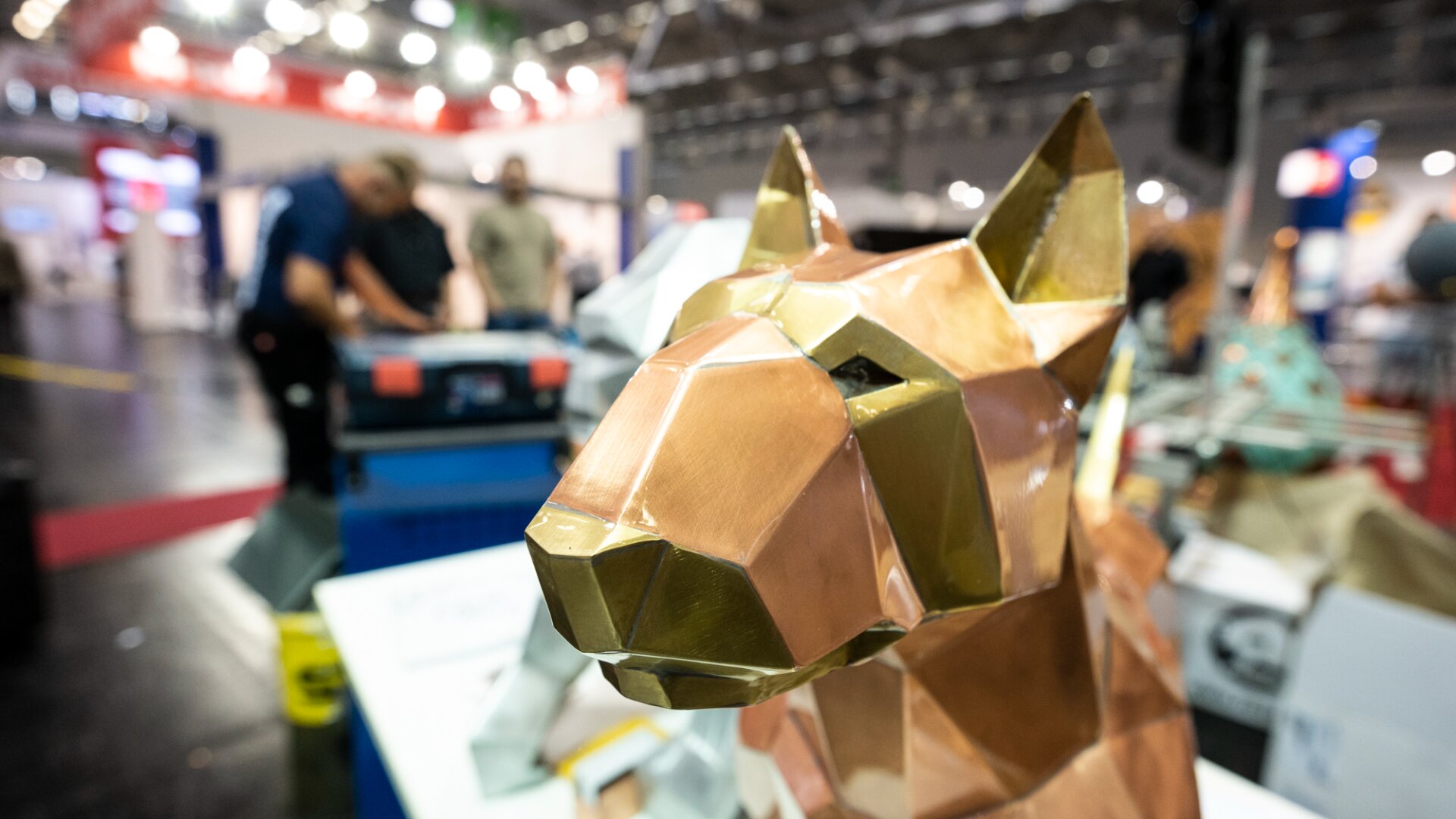 A close-up of a geometric, metallic dog sculpture is displayed indoors at a busy event, with people and equipment blurred in the background.