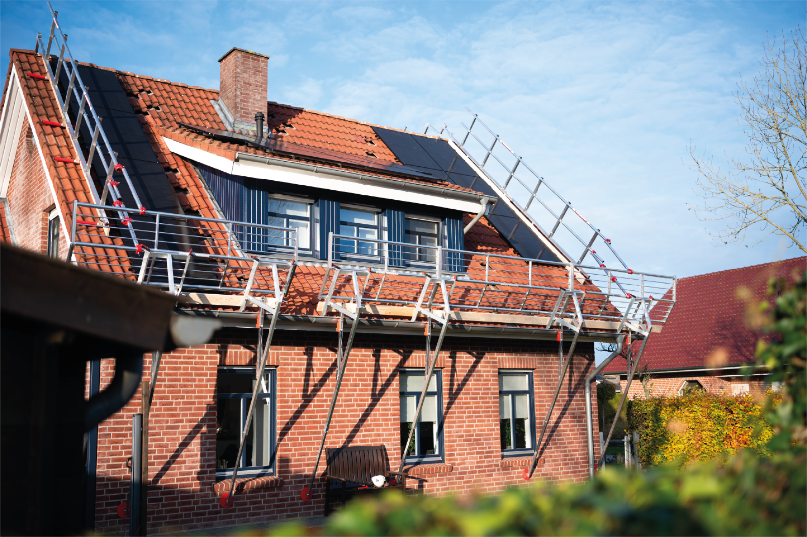 A brick house with scaffolding on the tiled roof, on which some tiles are missing. There are solar panels installed and a chimney is visible; the sky is clear and blue.