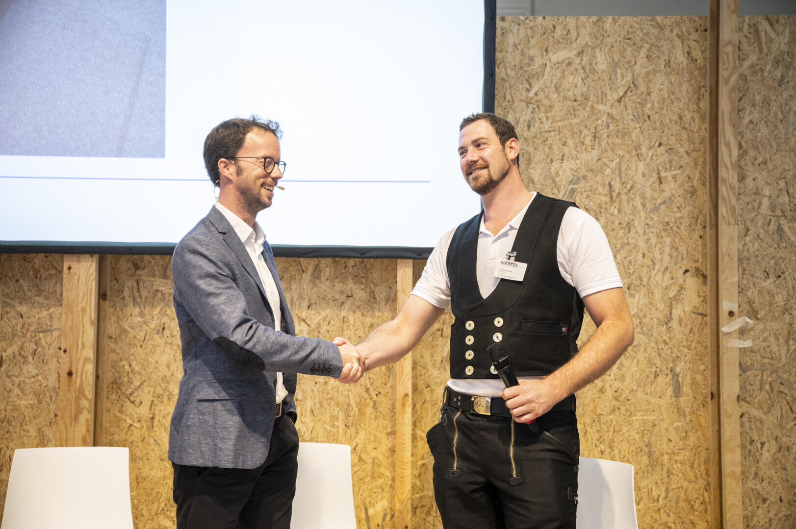 Zwei Männer schütteln sich lächelnd die Hände auf einer Bühne. Einer trägt einen blauen Blazer und eine Brille, der andere eine Weste und hält ein Mikrofon. Im Hintergrund sind eine Leinwand und eine Wand mit Holzstruktur zu sehen.