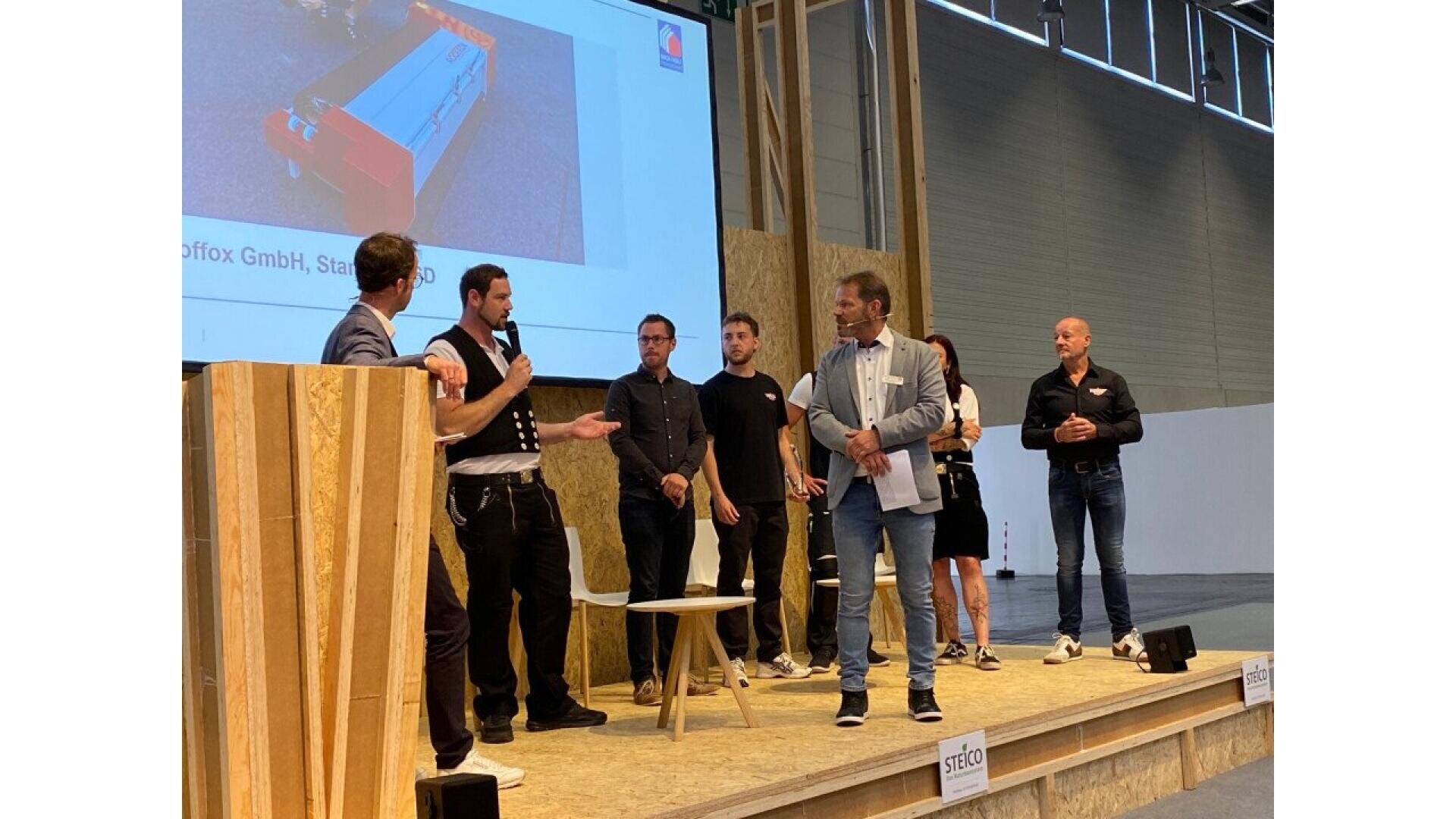 A group of seven people stand on a stage, some holding microphones and speaking to each other. A large screen with a product image is displayed behind them in a modern indoor event space.
