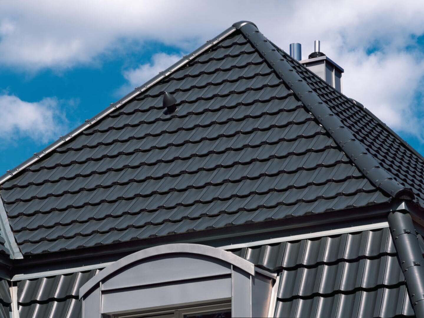 Eine Nahaufnahme eines mit dunkelgrauen Metalldachziegeln gedeckten Hausdachs mit einem kleinen Schornstein und einem Dachfenster vor einem blauen Himmel mit weißen Wolken.
