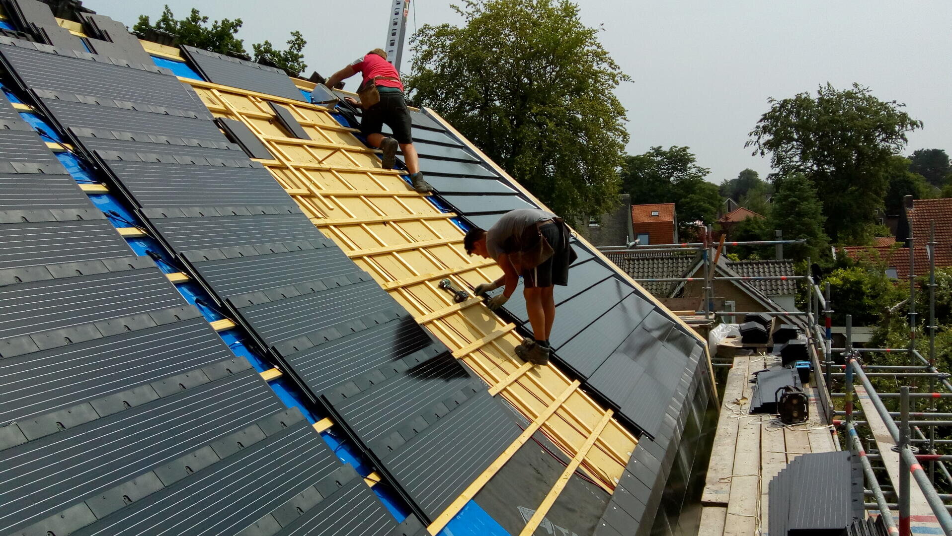 Two workers on scaffolding install solar modules on a pitched roof and attach the black modules to a wooden grid. On a clear day, trees and roofs can be seen in the background.