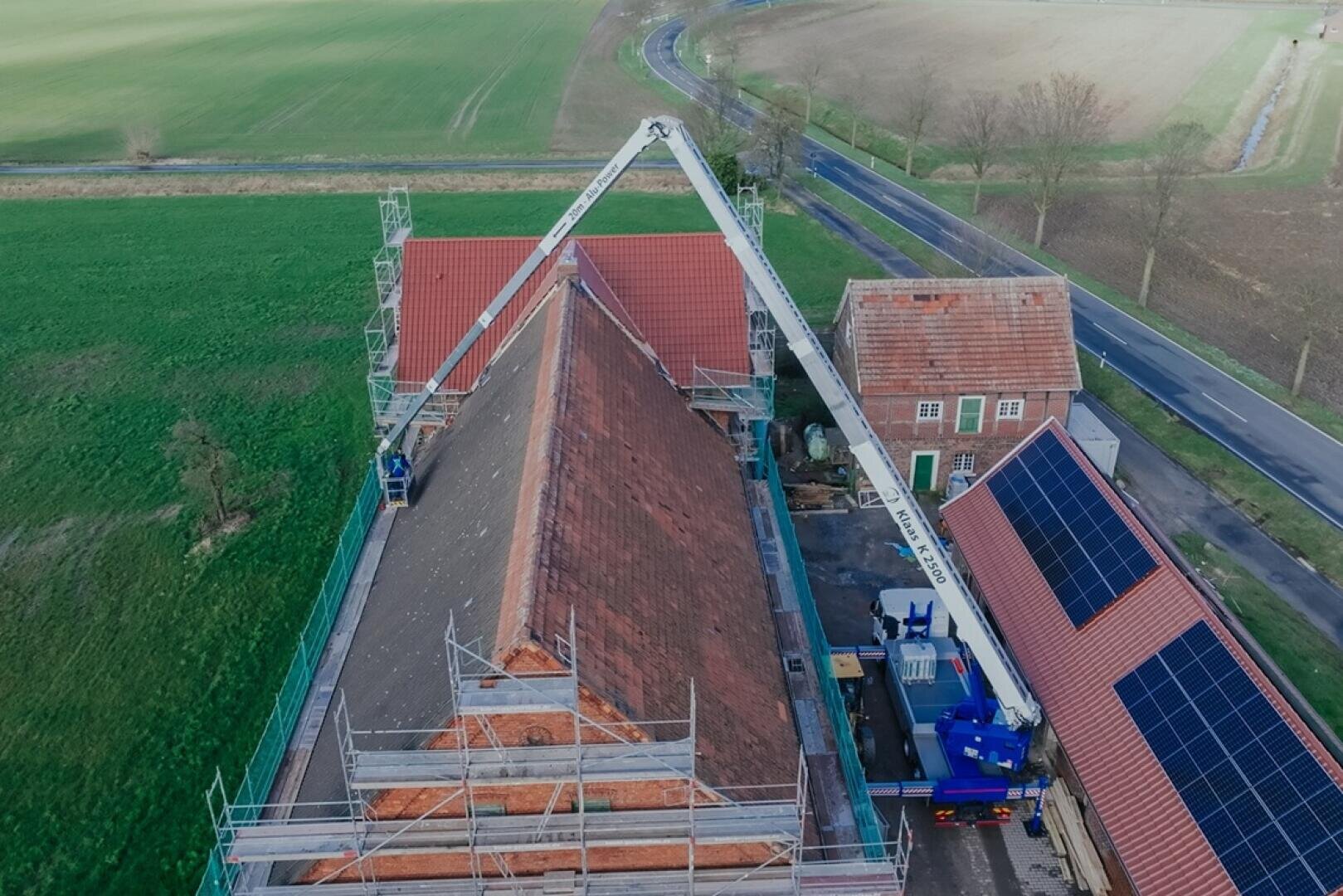 Luftaufnahme eines Gebäudes mit rotem Ziegeldach, das gerade renoviert wird, umgeben von einem Gerüst. Ein Kranwagen steht neben dem Gebäude, und auf einem angrenzenden Dach sind Solarzellen zu sehen. Im Hintergrund sind Felder und eine Straße zu sehen.