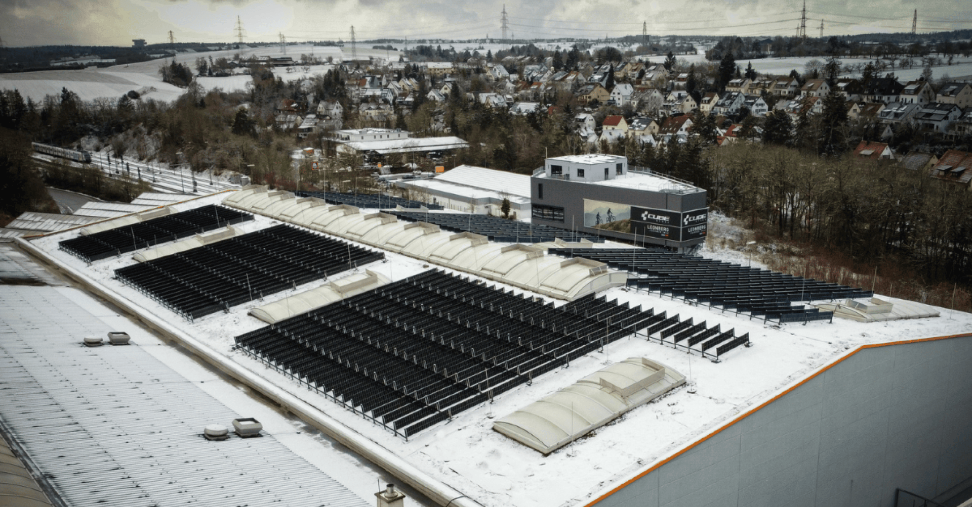 Ein großes Industriegebäude mit schneebedeckten Solarzellen auf dem Dach, umgeben von Bäumen und einem Wohngebiet im Winter. Die Dächer und der Boden in der Nähe sind ebenfalls mit Schnee bedeckt.