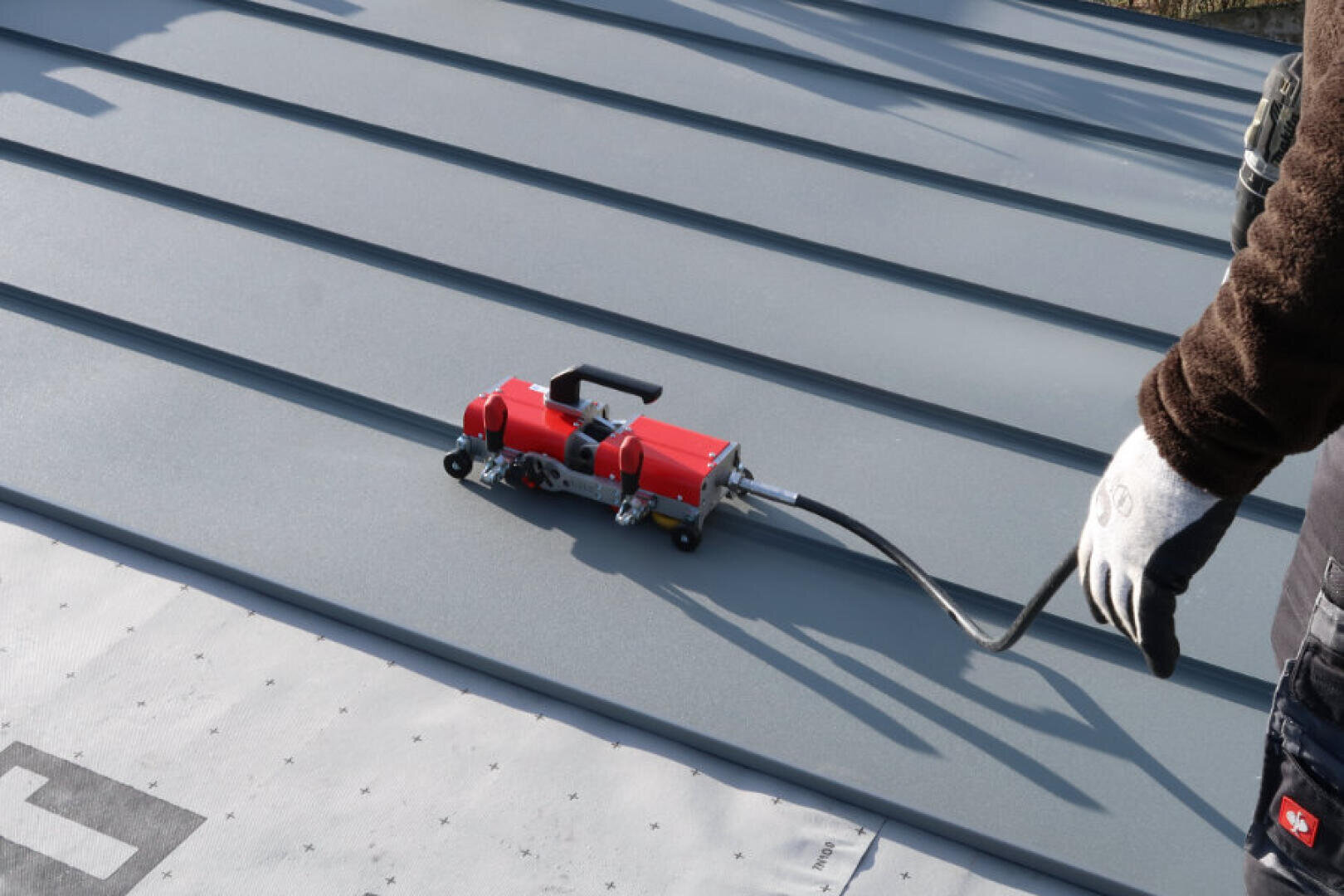 A person wearing gloves operates a red and black seam welder on a gray metal roof with a standing seam. The machine is connected to a cable and roofing material can be seen in the foreground.