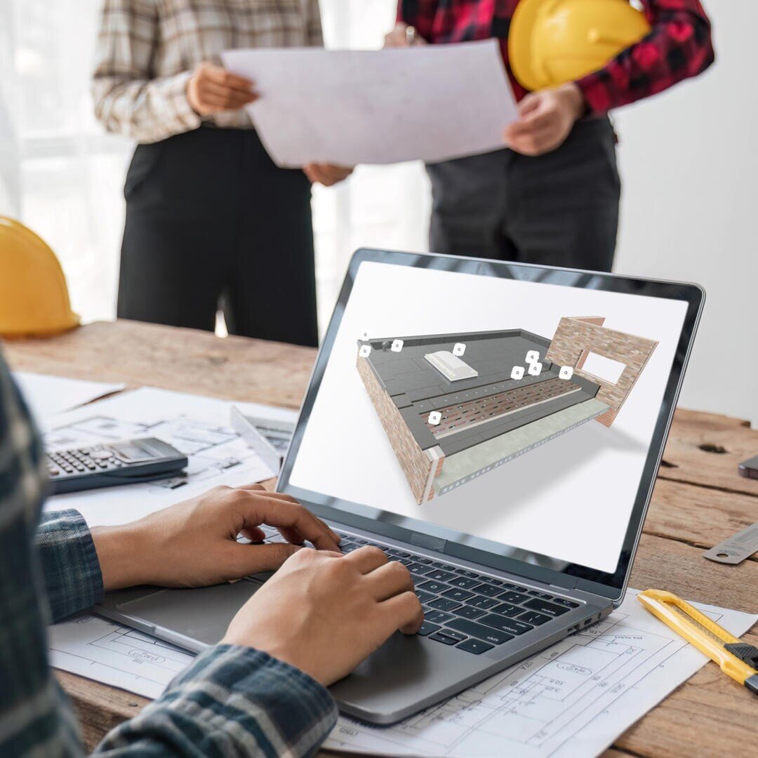 Eine Person verwendet einen Laptop mit einem 3D-Gebäudemodell auf dem Bildschirm, während zwei Personen in karierten Hemden und Schutzhelmen im Hintergrund auf einem Holztisch Baupläne prüfen.
