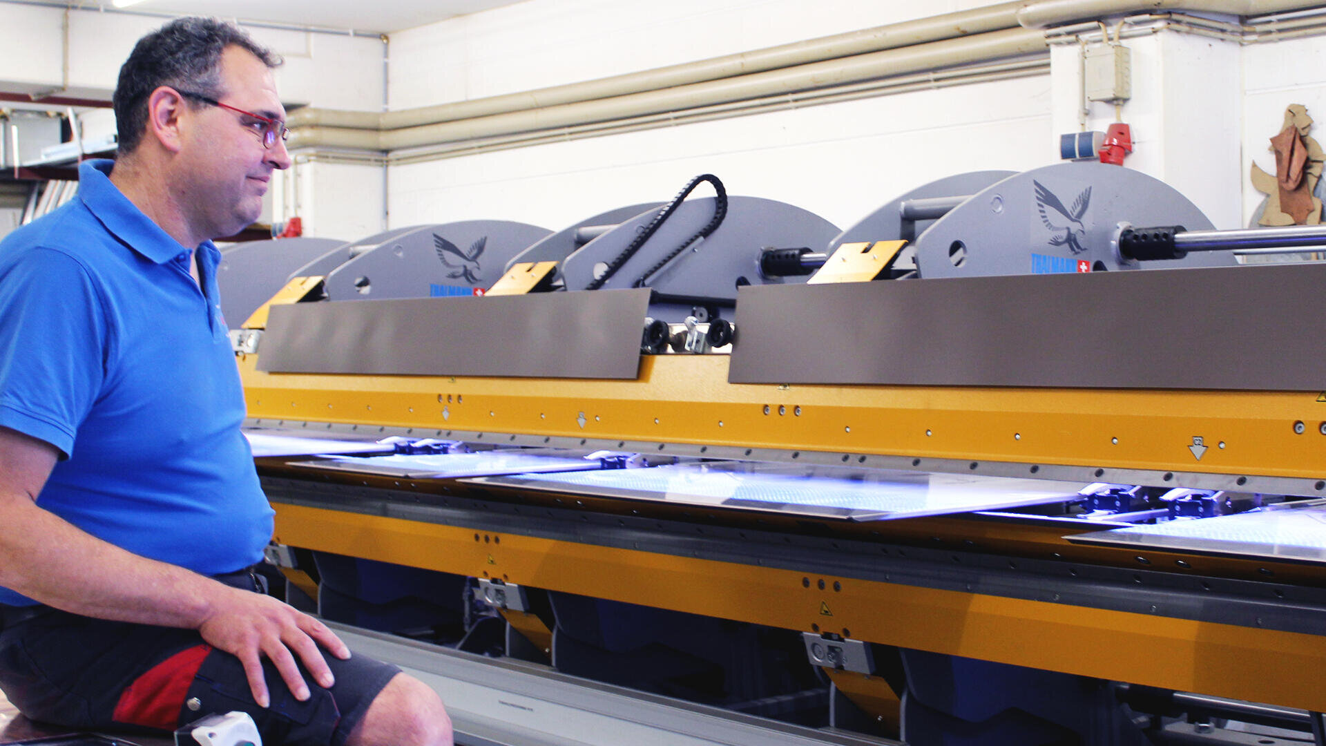 Ein Mann mit Brille und blauem Hemd bedient eine große gelb-graue Industriemaschine in einer hell erleuchteten Fabrik.