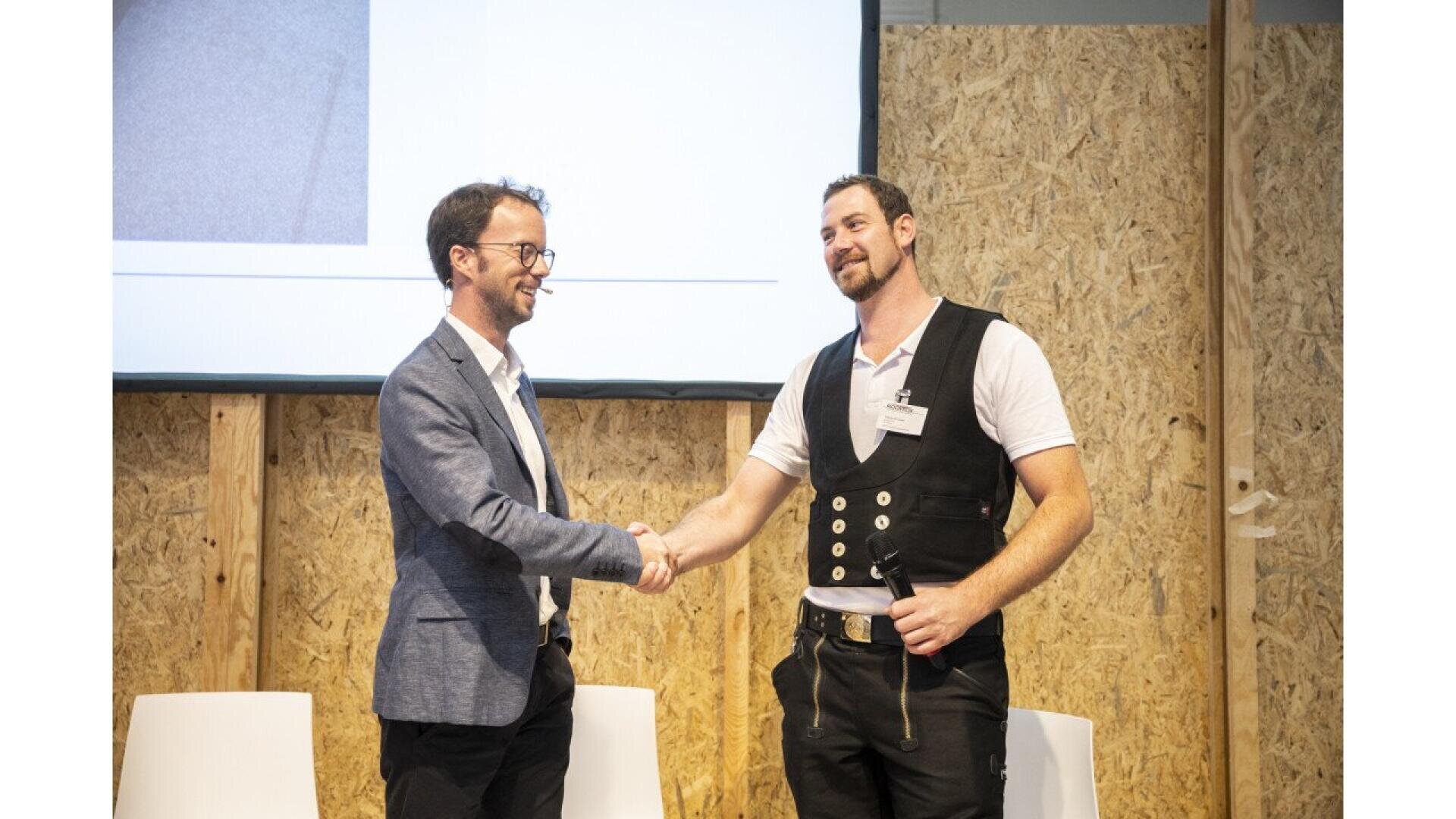 Two men are shaking hands and smiling on a stage. One wears a suit jacket and glasses; the other wears a white shirt with a black vest and a name tag. A screen and wooden wall are visible in the background.