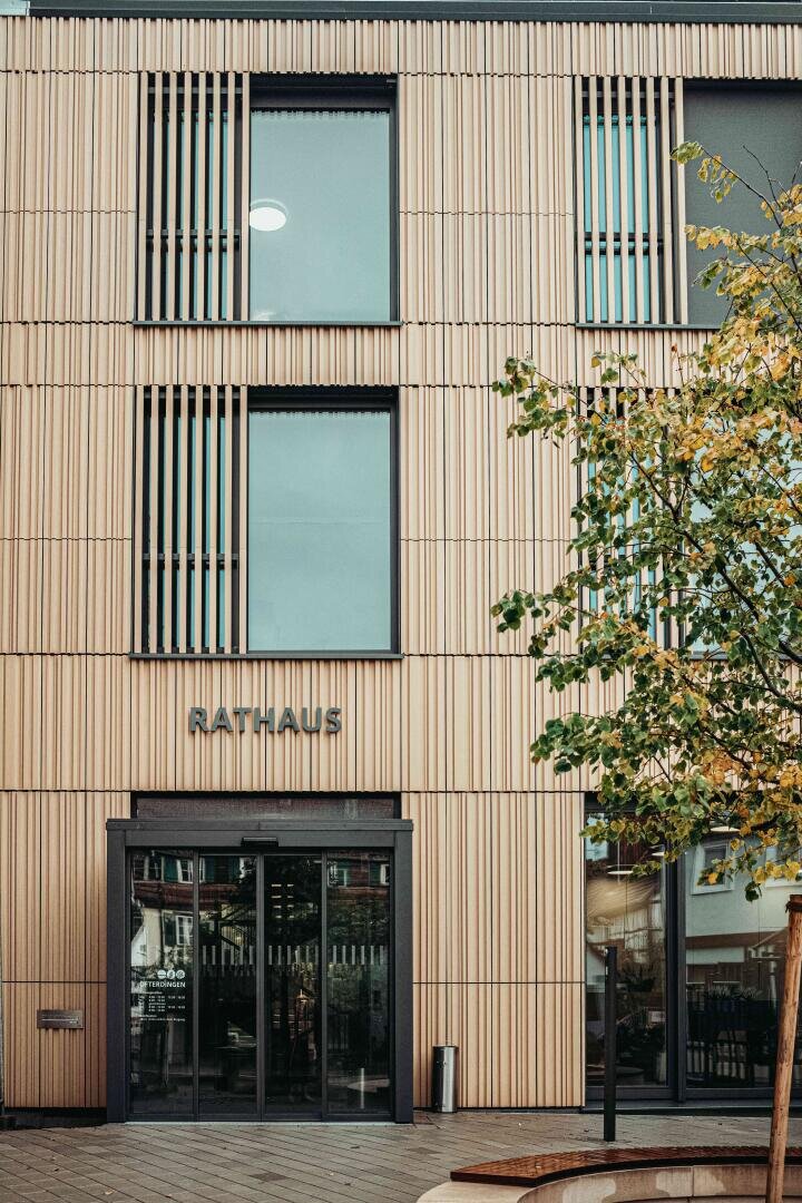 Modern building with vertical wooden slats, large windows and a glass entrance. The word Rathaus is written above the door. In the foreground on the right is a tree with green leaves.