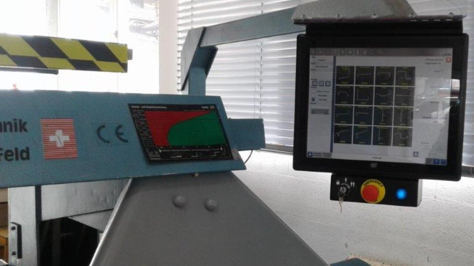 A close-up of industrial machinery features a control panel with graphs on a screen and another monitor displaying multiple graphs and buttons. The machine has safety markings and is situated near a window with blinds.