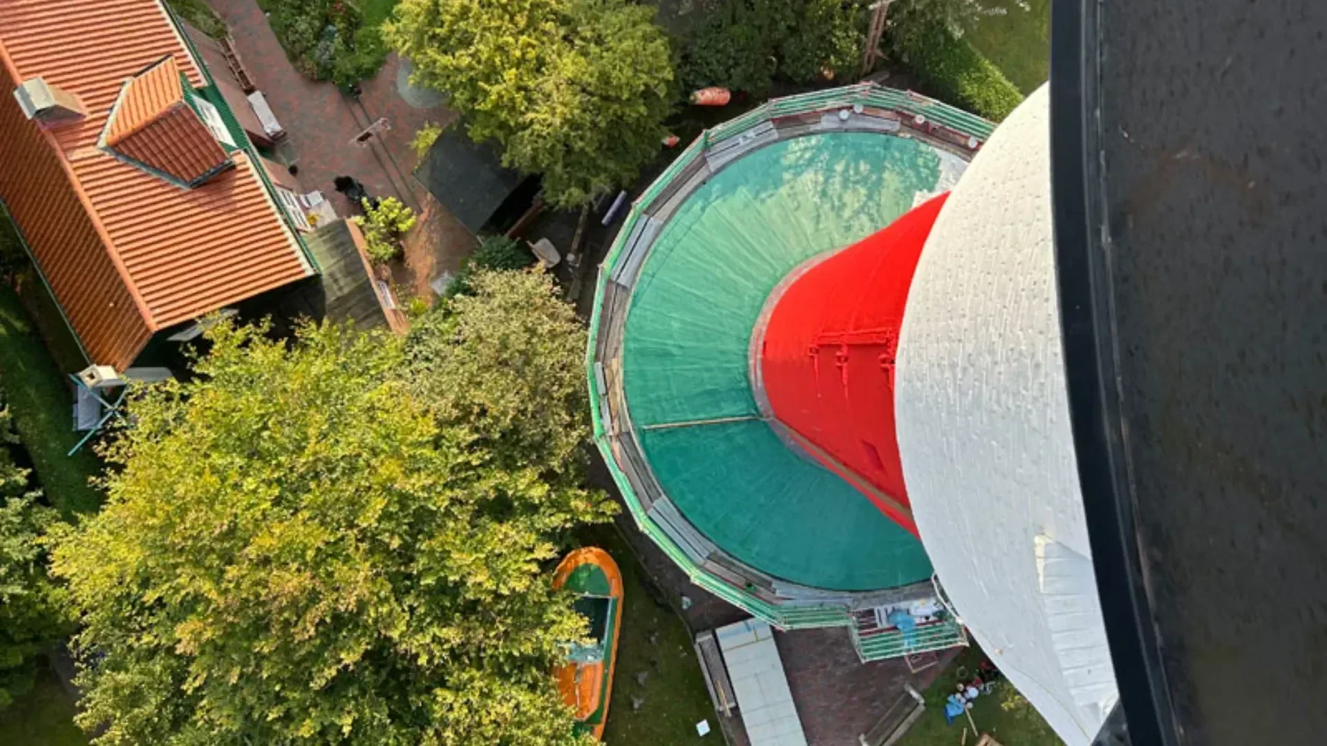 Luftaufnahme mit Blick direkt nach unten auf die Spitze eines rot-weißen Leuchtturms, umgeben von grünen Bäumen, Gebäuden und Gehwegen im Freien.