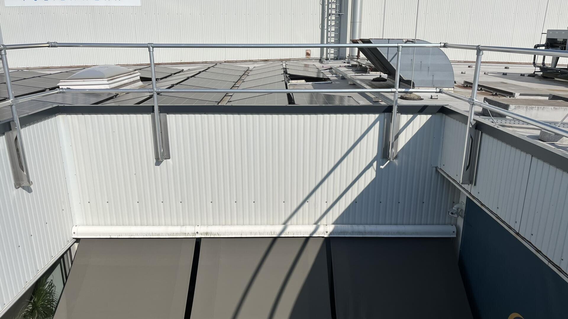 Ein Dachbereich mit Metallgeländern, Sonnenkollektoren und HVAC-Kanälen. Die Schatten des Geländers fallen auf die weiße Wellblechwand und die grauen Markisen darunter.