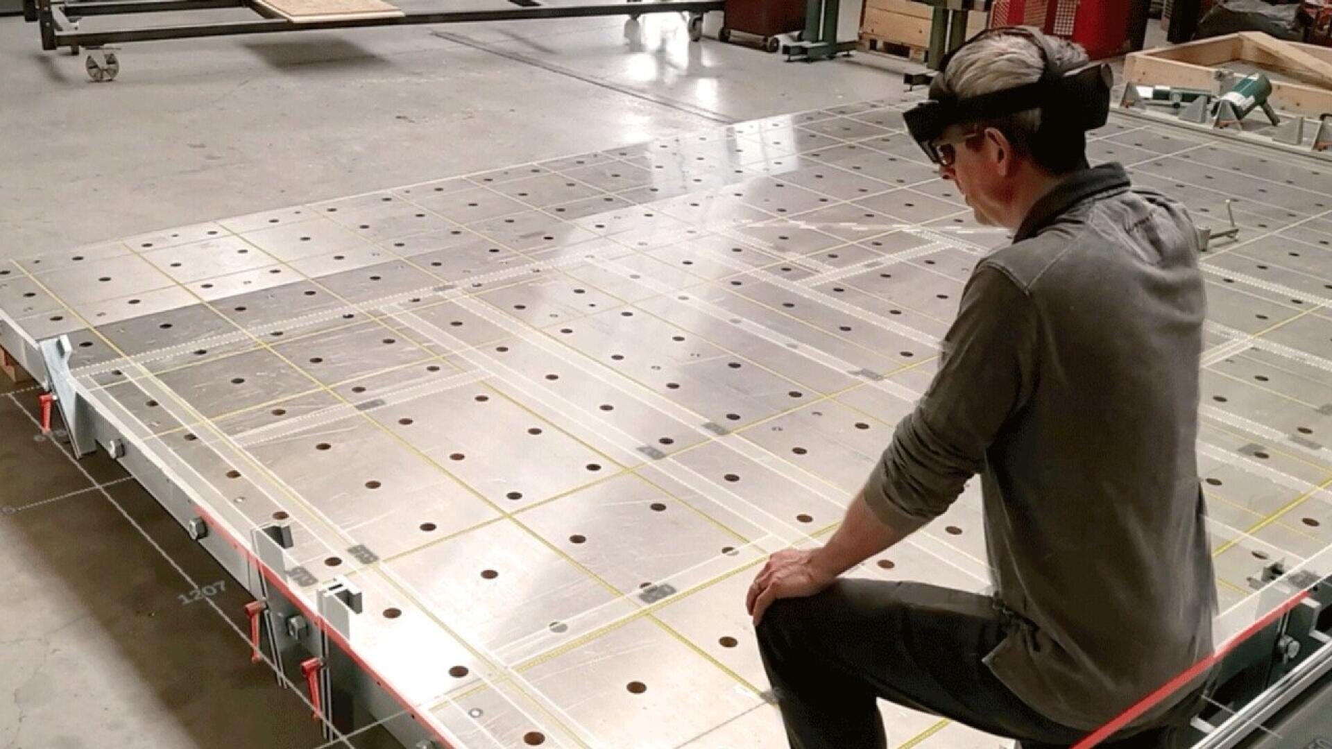 A person wearing augmented reality glasses kneels beside a large metal surface with evenly spaced holes, in an industrial or workshop setting. Yellow lines and grids are projected across the surface.