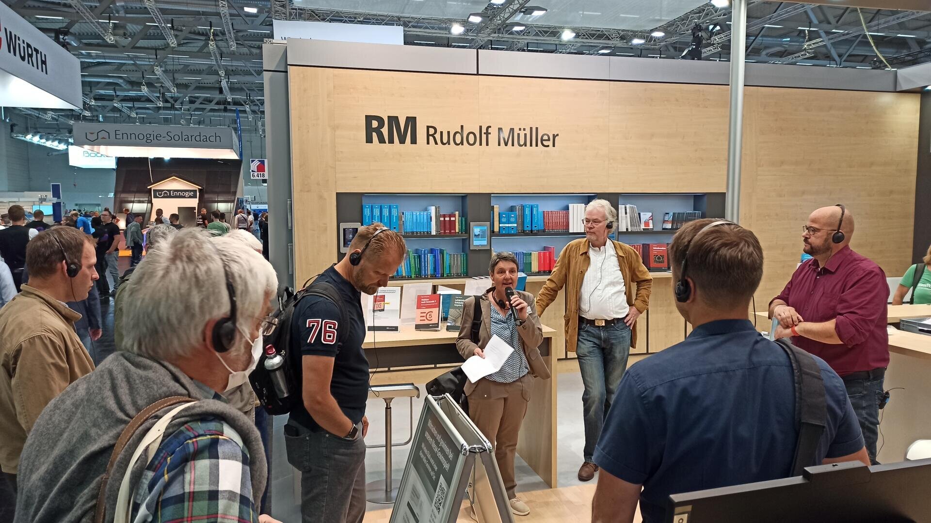 A group of people wearing headsets listen to a woman speaking with a microphone at the Rudolf Müller booth at a trade fair, surrounded by books and display materials.