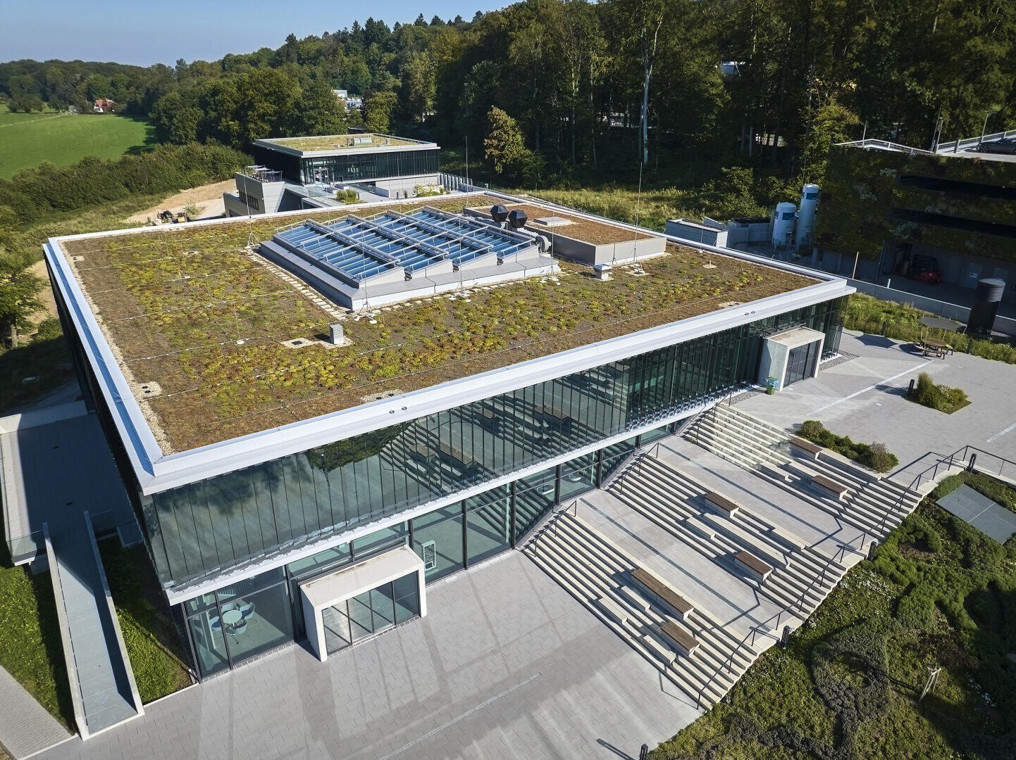 Ein modernes Gebäude mit großen Glasfenstern und einem begrünten Dach mit Pflanzen, umgeben von Bäumen und begrünten Wegen, an einem sonnigen Tag von oben betrachtet.