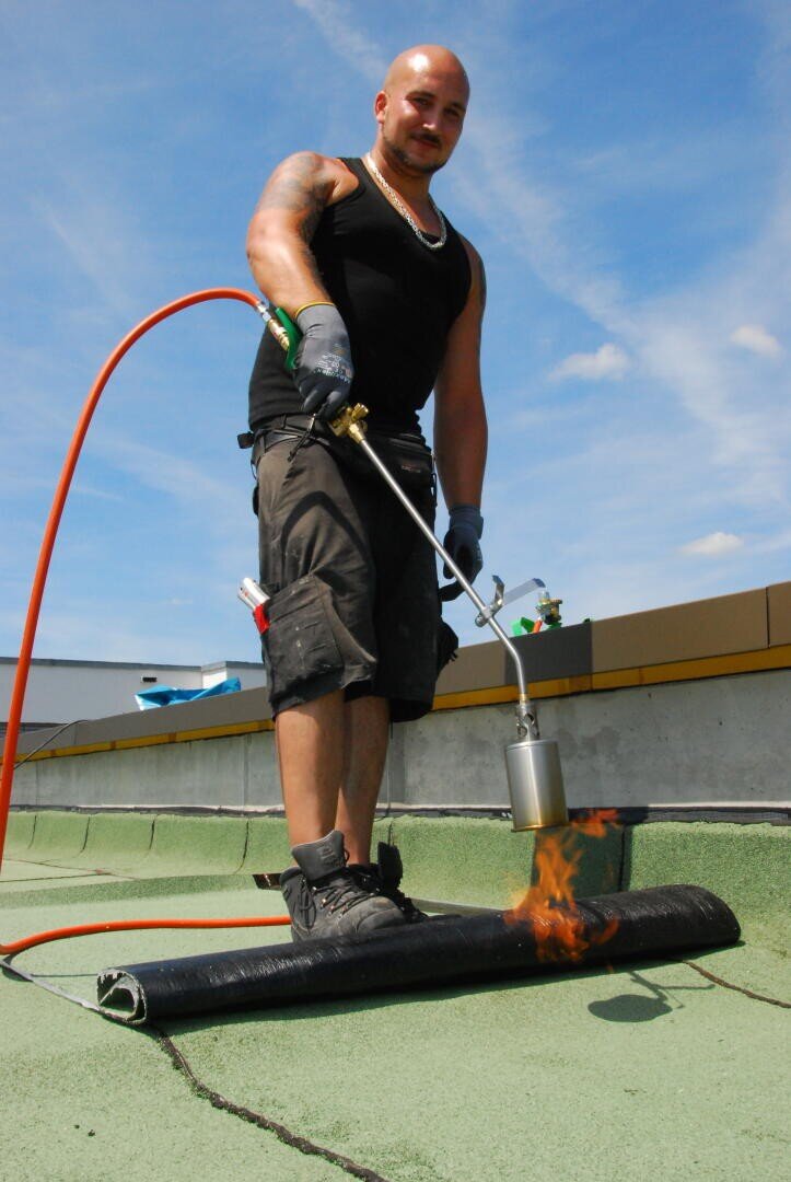 Ein Mann in schwarzem Tanktop und Shorts trägt unter blauem Himmel mit einem Gasbrenner Dachmaterial auf einem Flachdach auf. Ein orangefarbener Schlauch ist mit dem Brenner verbunden, und in der Nähe des Materials ist eine Flamme sichtbar.