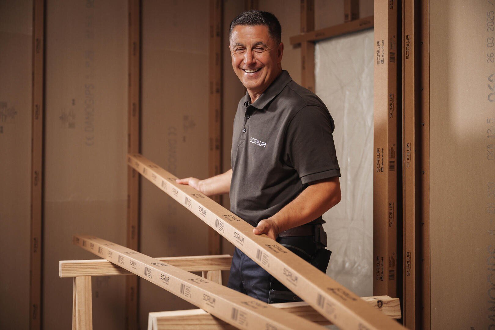 A smiling man in a gray polo shirt holds two long wooden planks into a building under construction with exposed beams and cardboard insulation panels.