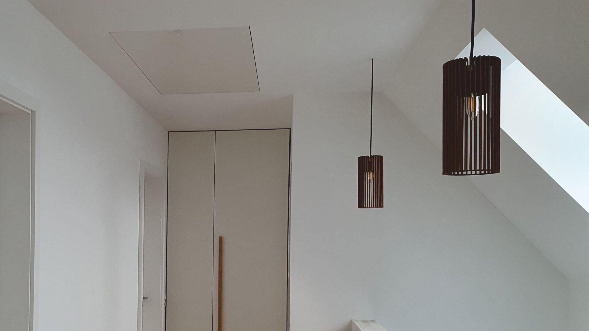 Ein minimalistisches Interieur mit weißen Wänden, einer geschlossenen Tür, einem Einbauschrank, zwei modernen Pendelleuchten mit dunklen Lamellenschirmen und einem großen Oberlicht, das natürliches Licht hereinlässt.