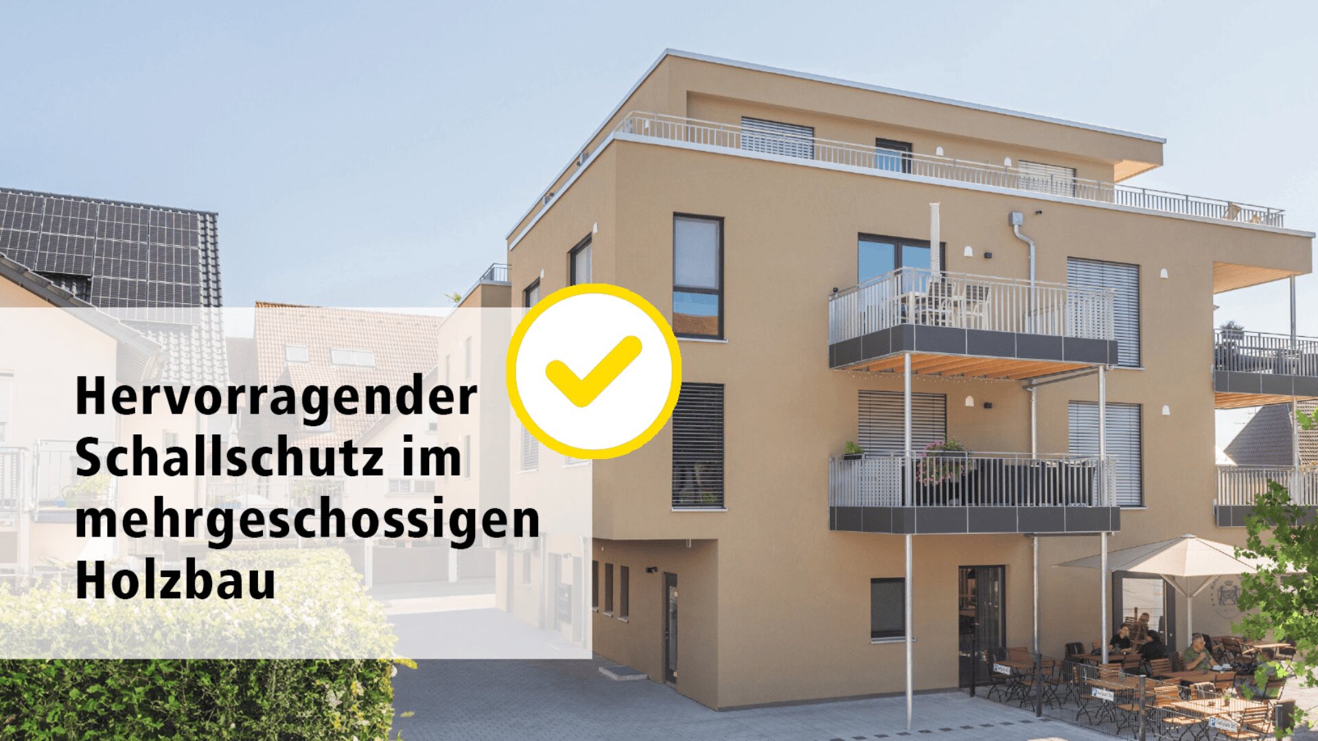 Modernes dreistöckiges Mehrfamilienhaus mit Balkonen. Neben einem gelben Häkchen steht „Hervorragender Schallschutz im mehrgeschossigen Holzbau“. Das Gebäude ist von Grünflächen und einer Terrasse umgeben.