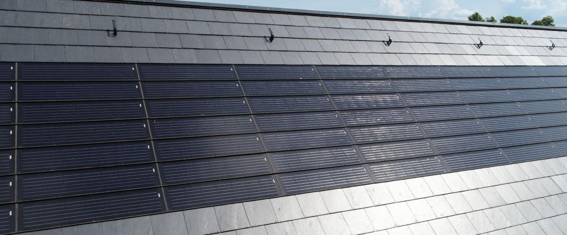 Nahaufnahme eines Schrägdachs mit integrierten Solarmodulen, umgeben von grauen Dachziegeln, unter einem teilweise bewölkten Himmel.