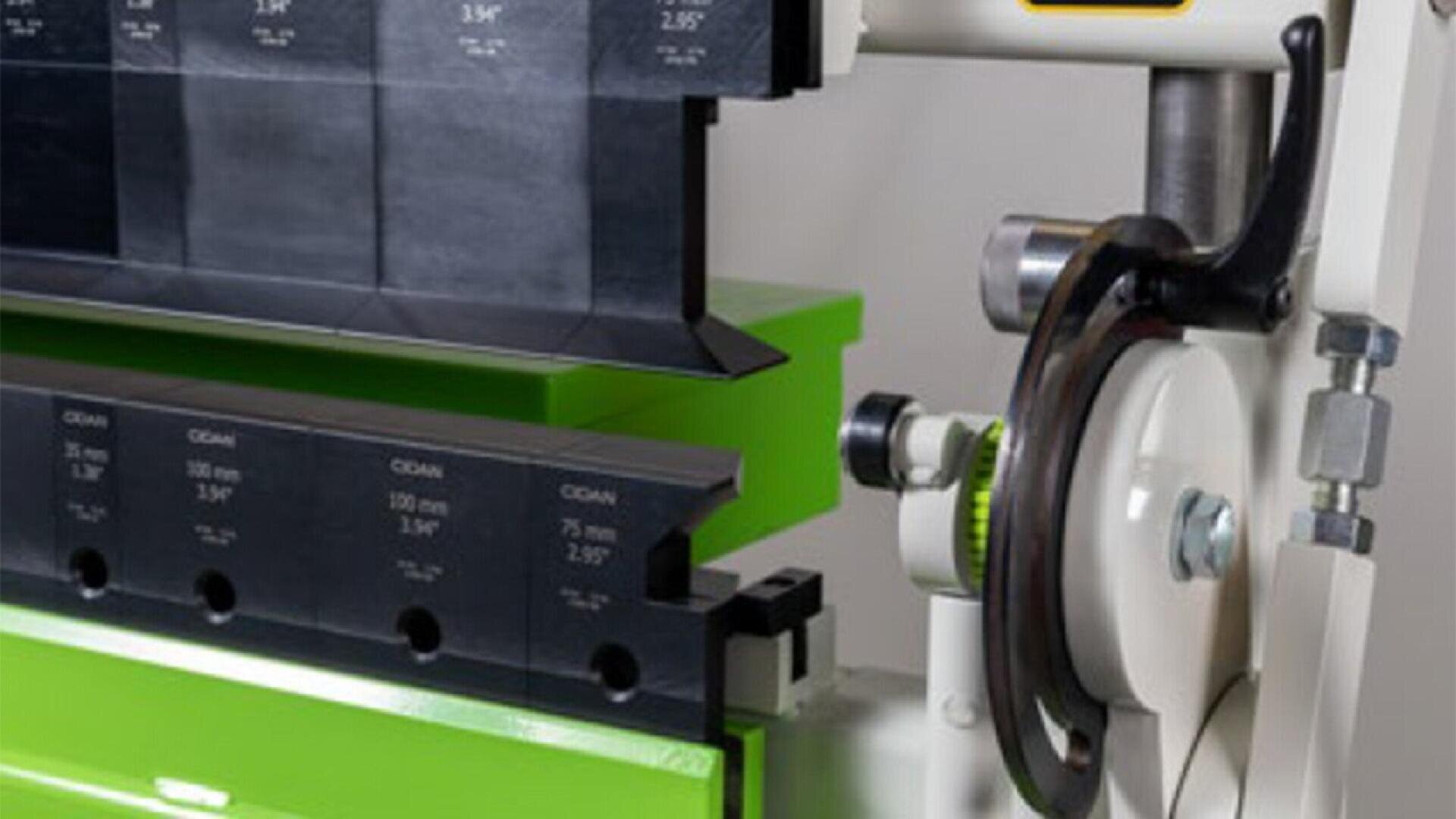 Close-up of a metalworking machine with black and green components, including measurement markings and a circular handwheel, likely used for bending or shaping metal sheets.