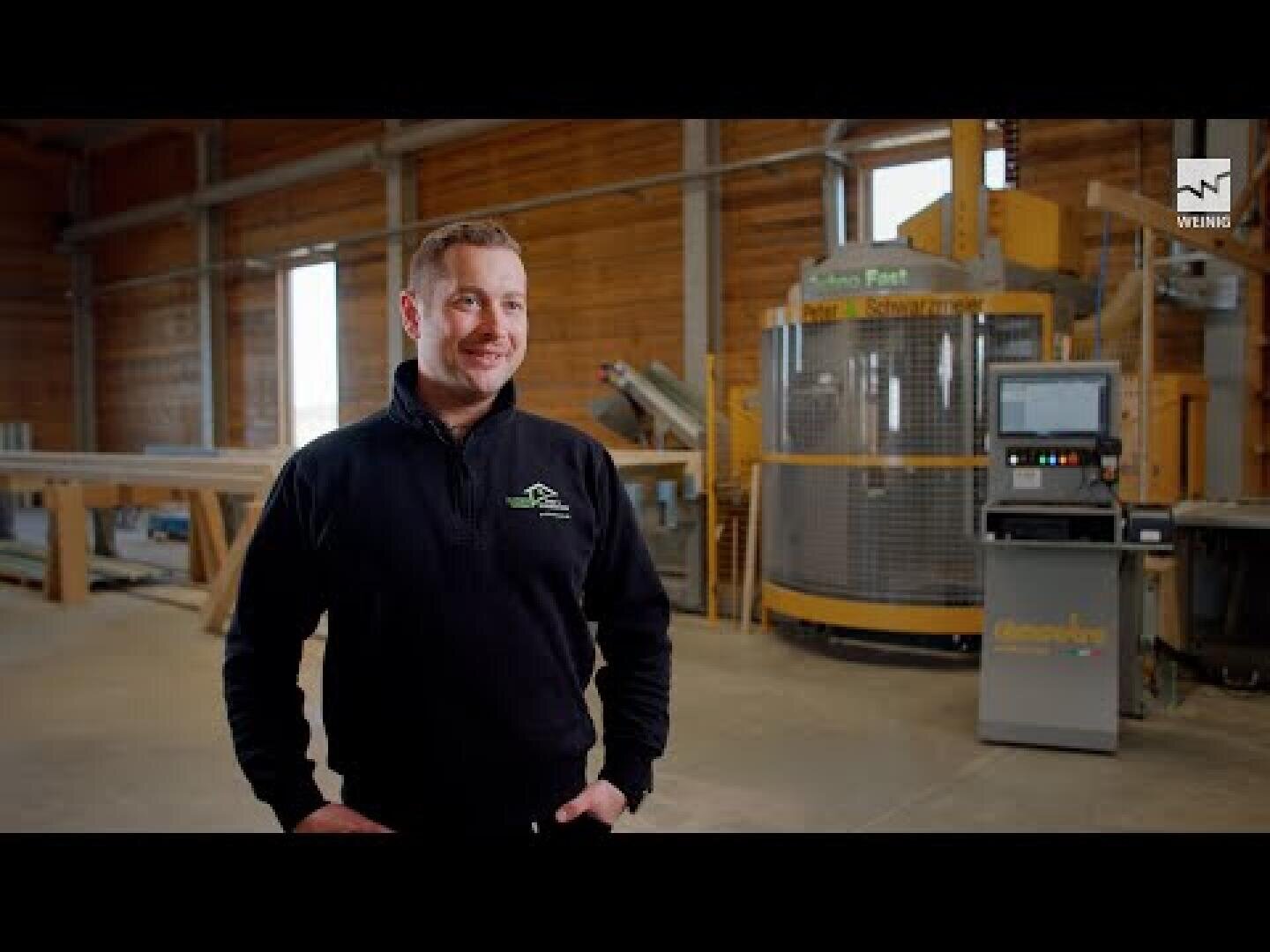 A man in a dark sweater stands smiling in a spacious, well-lit industrial hall with wooden walls and large machines in the background.