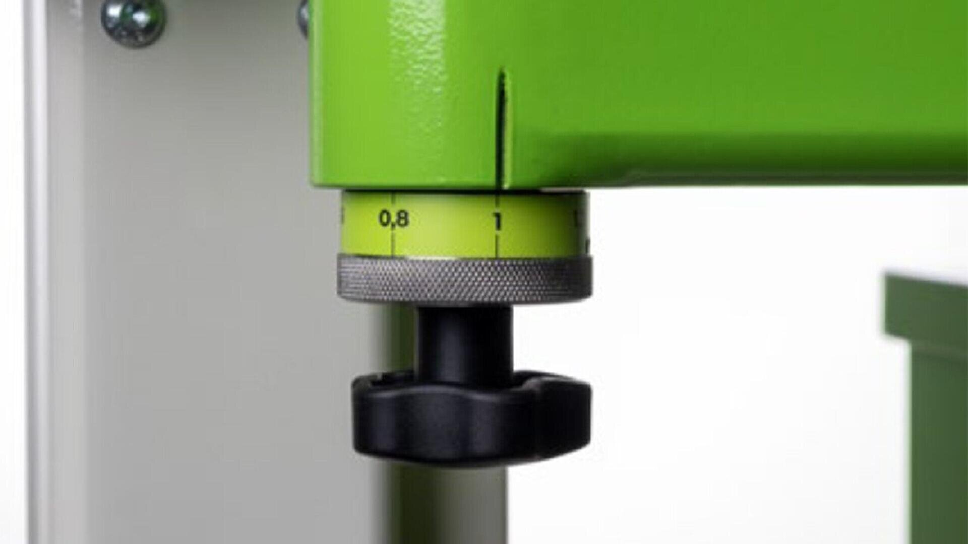Close-up of a green metal machine with a black adjustment knob and a yellow dial marked with numbers from 0.8 to 1, likely indicating a measurement or setting. The background is out of focus.
