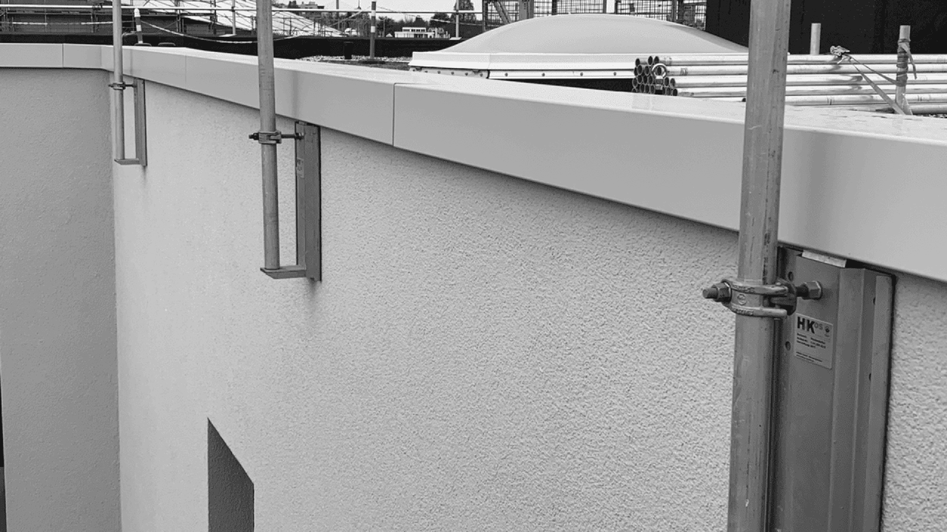 A close-up view of a building’s exterior wall with scaffolding and construction materials on the rooftop, shown in black and white. The wall has a textured finish and there are metal brackets attached.