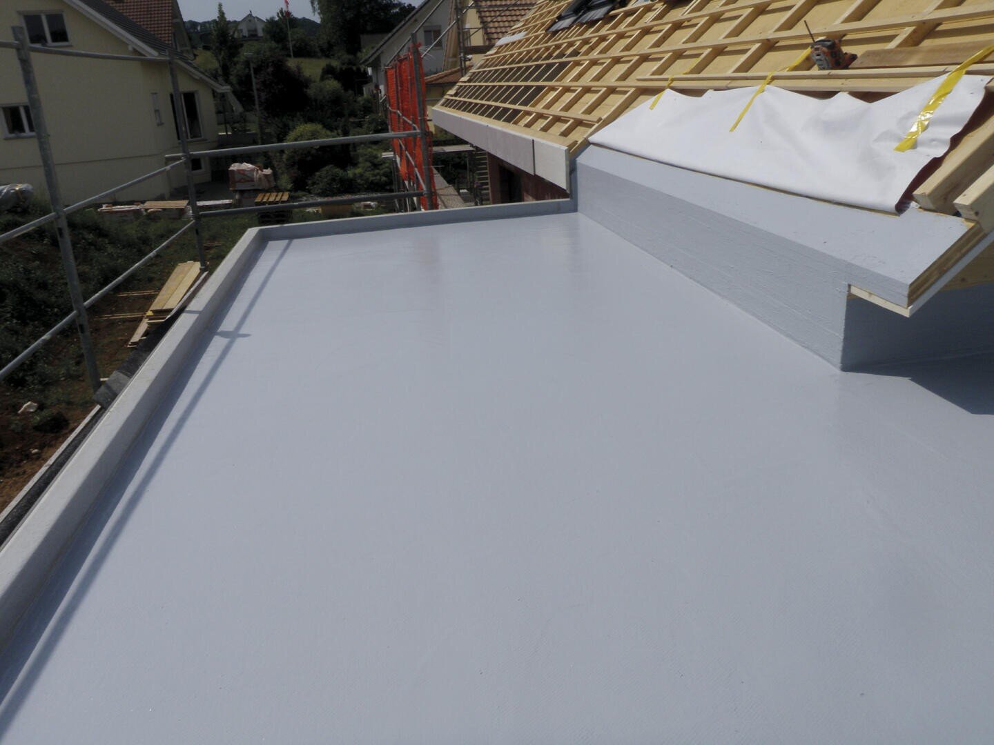 Ein Flachdach, das mit einer glatten, hellgrauen, wasserdichten Beschichtung bedeckt ist, neben einem im Bau befindlichen Schrägdach mit Holzbalken und teilweise installiertem Dachmaterial. Im Hintergrund sind Häuser und Grünflächen zu sehen.