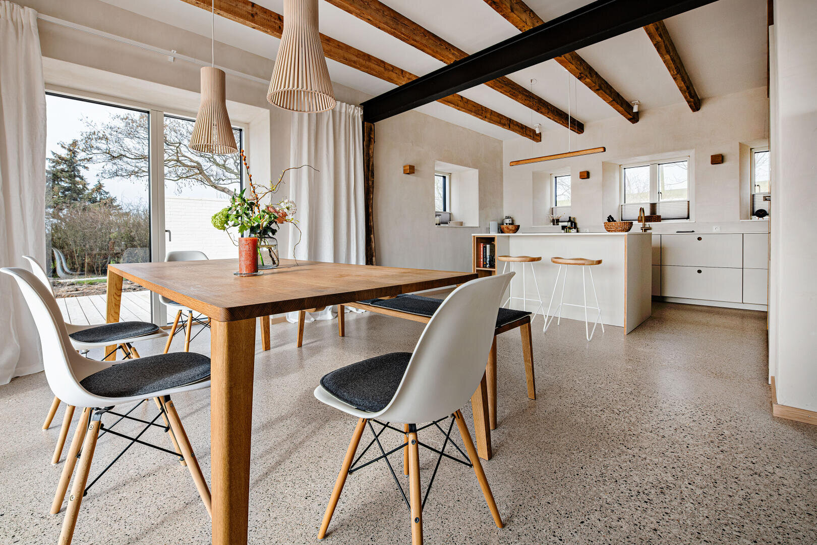 A modern dining and kitchen area with wooden beams, large windows, a wooden table with white chairs, pendant lights, minimalist white cabinets and a view of the trees outside.